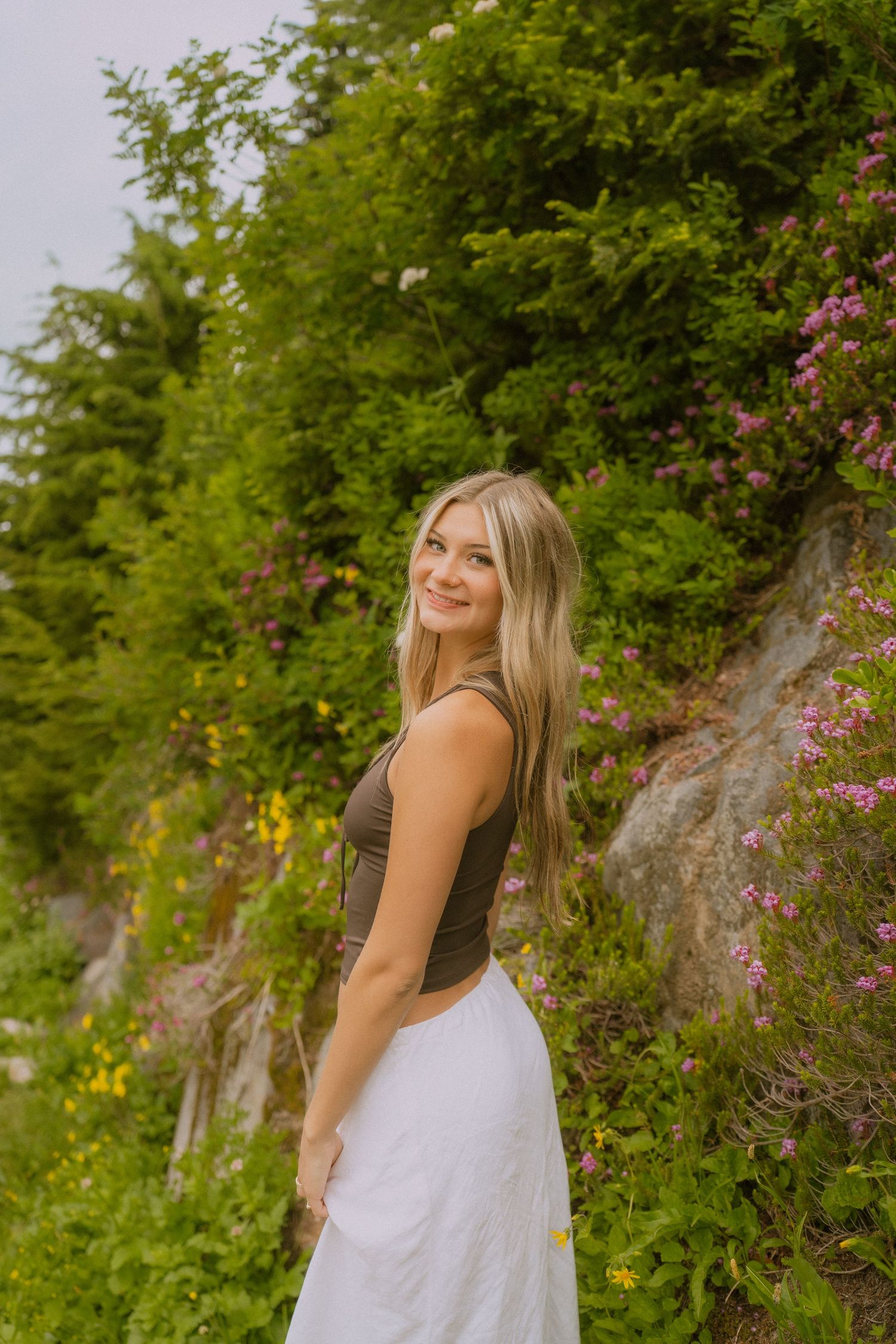 A woman in a white dress poses in front of lush greenery and pink flowers on a summer day.