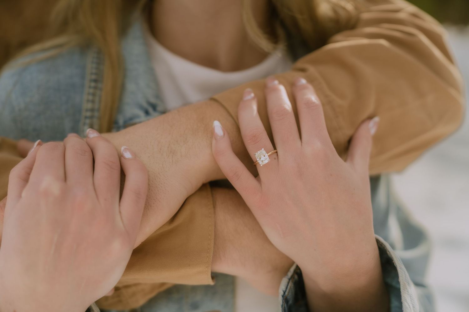 Close-up view of hands wearing a sparkly engagement ring against denim and beige clothing.