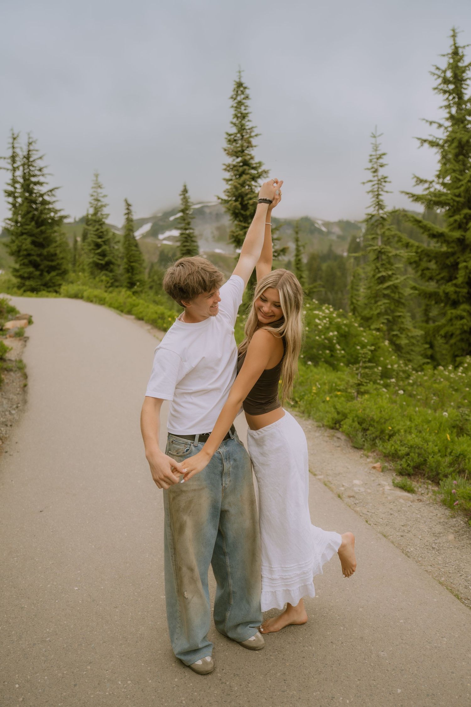 A couple dances together on a mountain path surrounded by evergreen trees and scenic views.