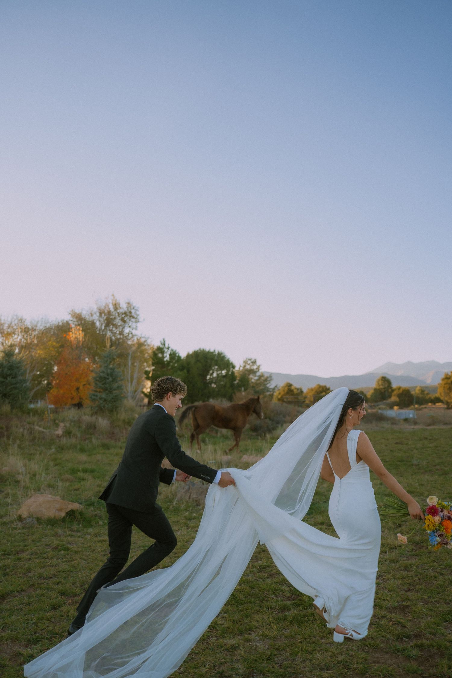 A couple in wedding attire dances playfully in a field during autumn with the bride's veil flowing in the breeze.