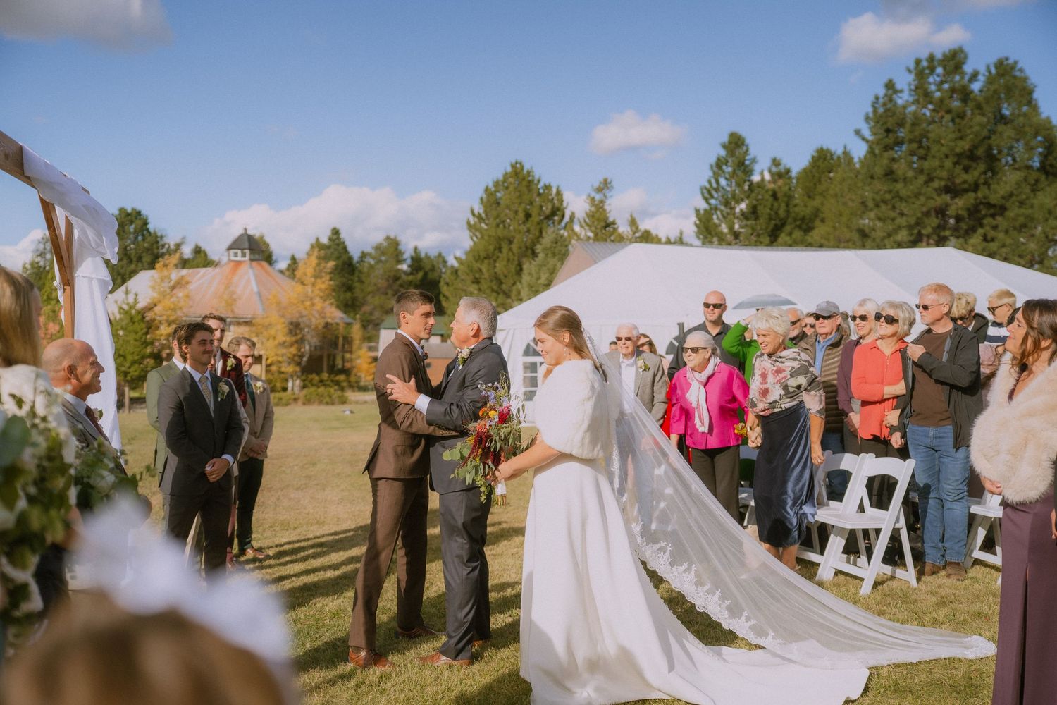 A wedding ceremony takes place outdoors on a sunny autumn day with guests seated on white chairs.