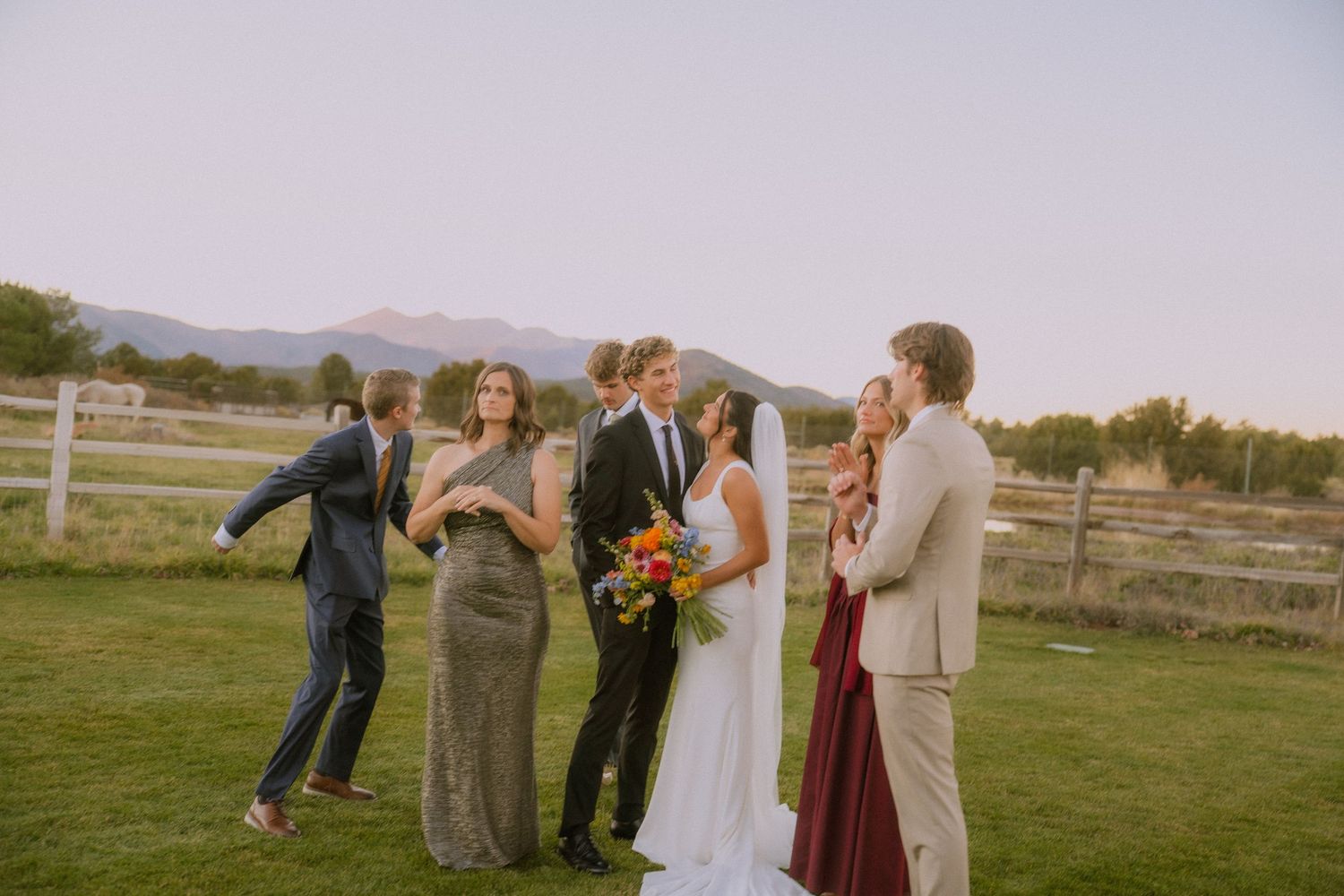 Wedding party members celebrating and dancing on grass with mountains in background.