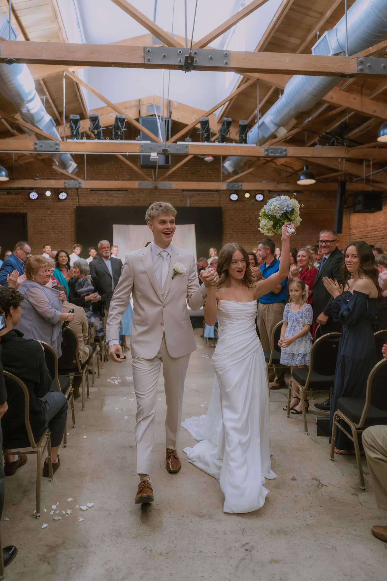 Wedding guests line up in a rustic indoor venue with exposed wooden beams and industrial lighting.