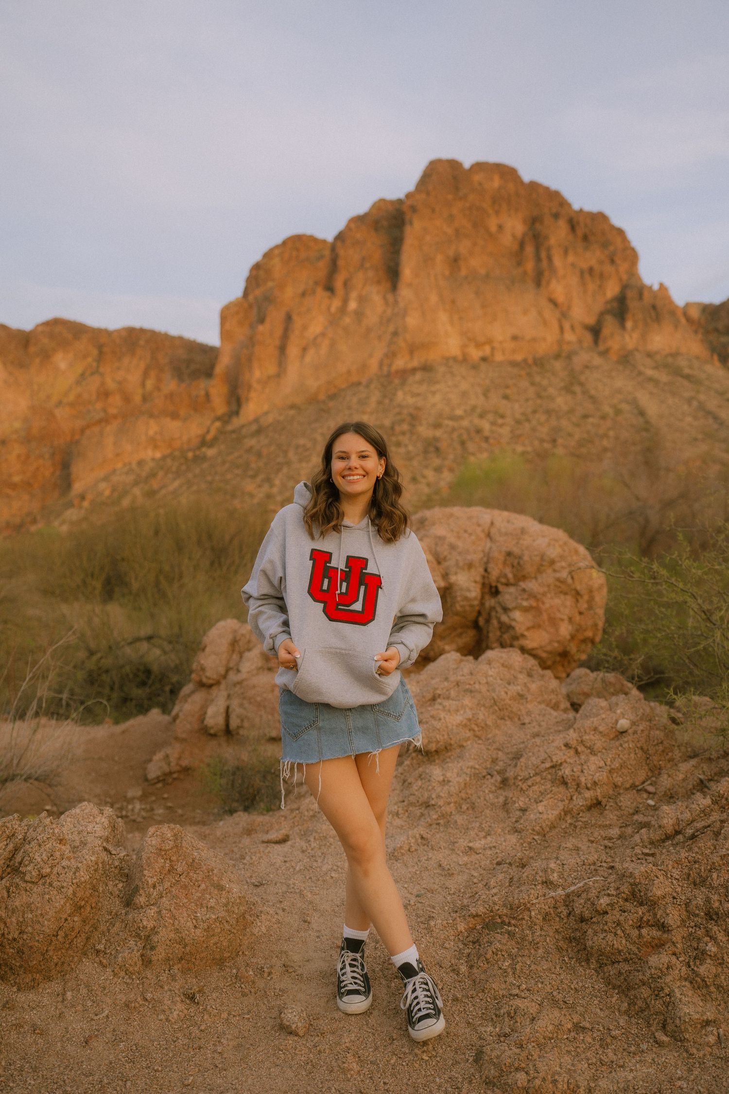 Someone wearing a University of Utah sweatshirt poses on rocky terrain with scenic desert cliffs in the background at sunset.