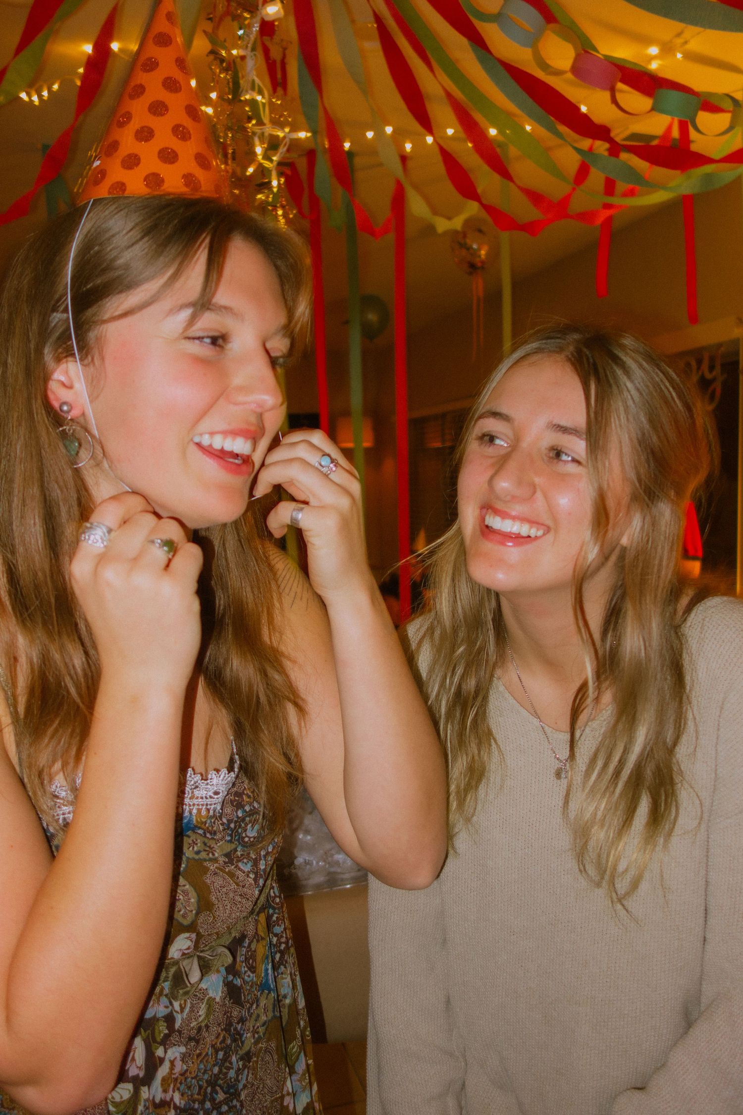 Two friends laugh and smile together under festive red and gold party decorations at a celebration.