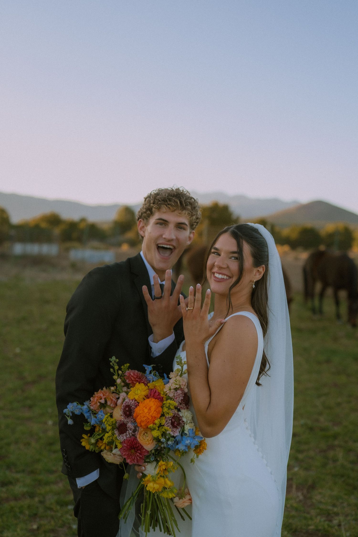 Newlyweds share a joyful laugh while holding a vibrant wildflower bouquet in an outdoor setting.