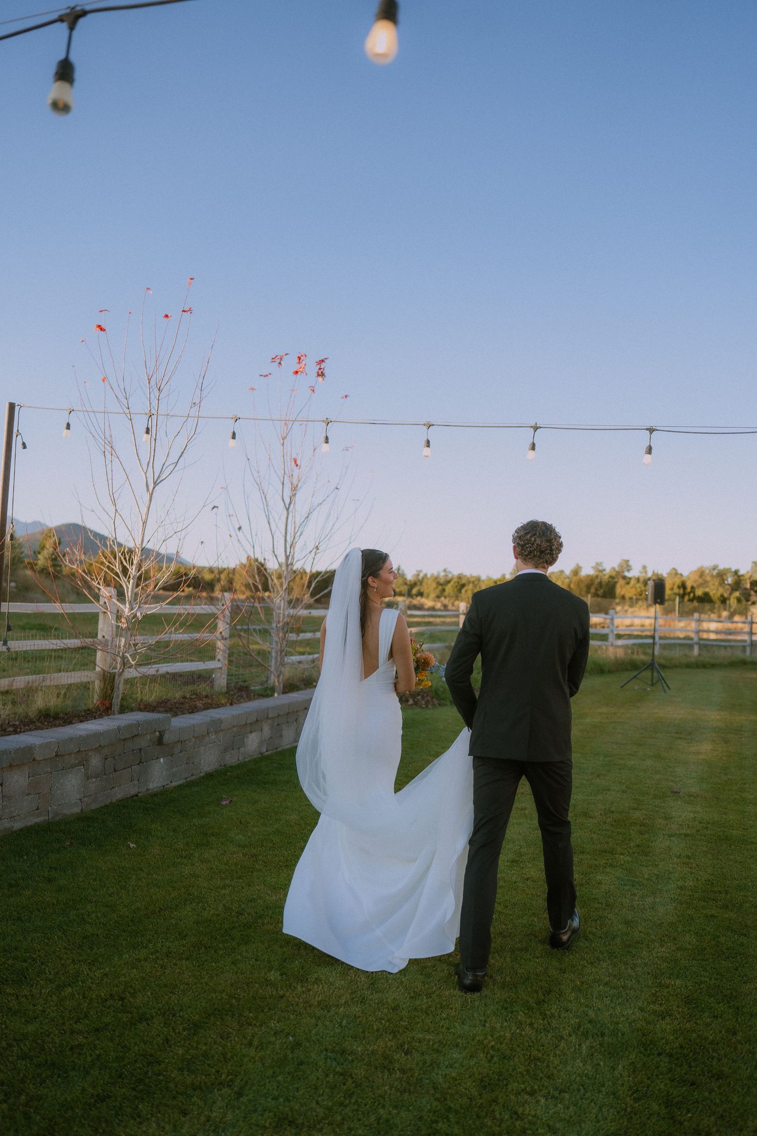 A couple in wedding attire standing together under string lights at dusk with mountains in the background.