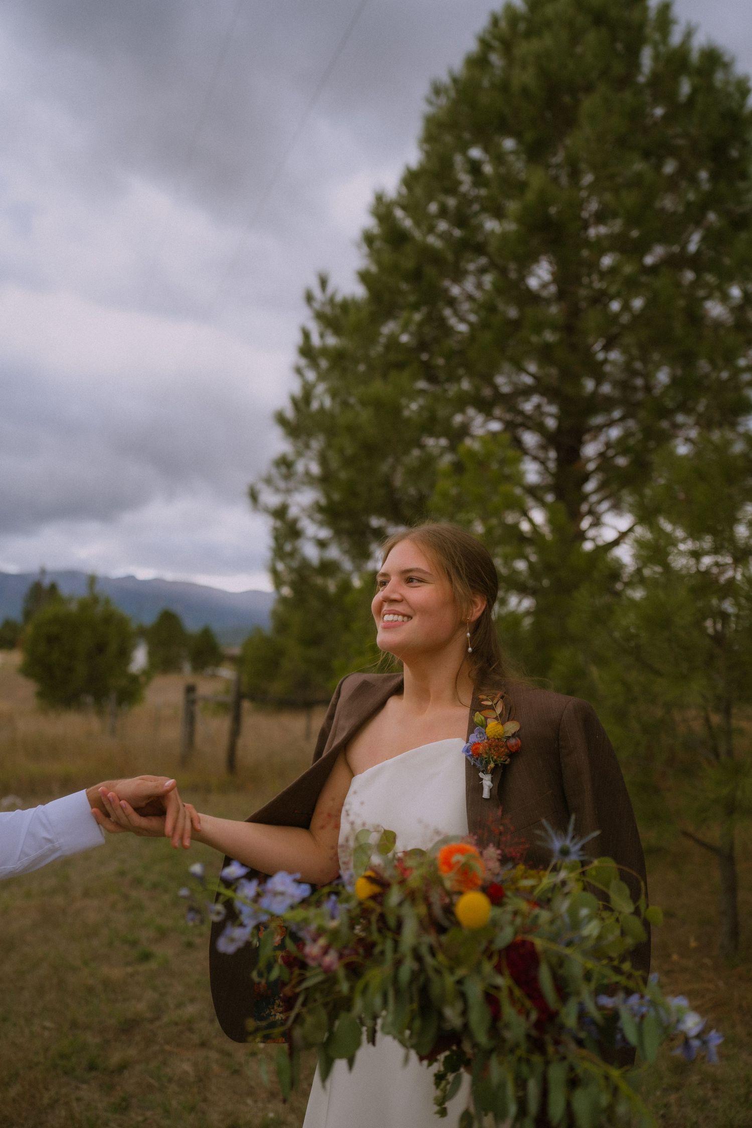 A bride in a white dress laughs joyfully while holding a colorful autumn bouquet in an outdoor field setting.
