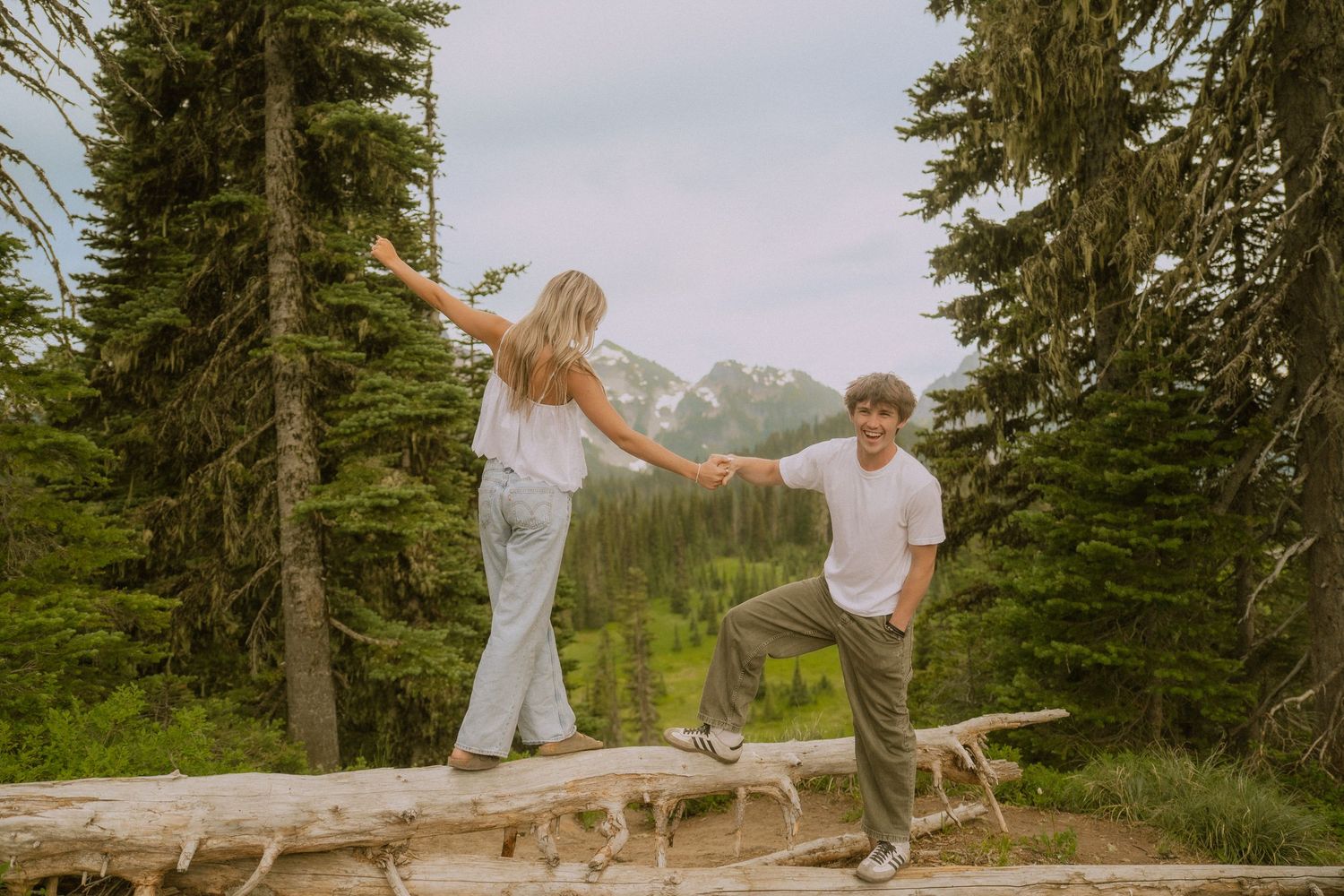 Couple balancing on a fallen log in a mountain forest setting during sunset.
