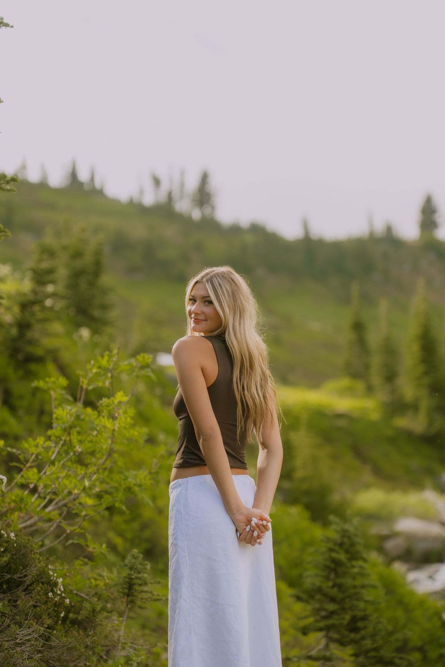 A person in a light blue dress stands among evergreen trees and mountain scenery on a summer day.