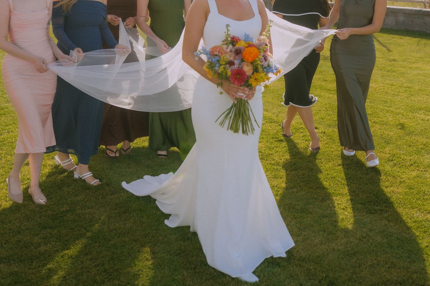 A flowing white wedding dress with a colorful bouquet catches sunlight on a grassy lawn during an outdoor ceremony.