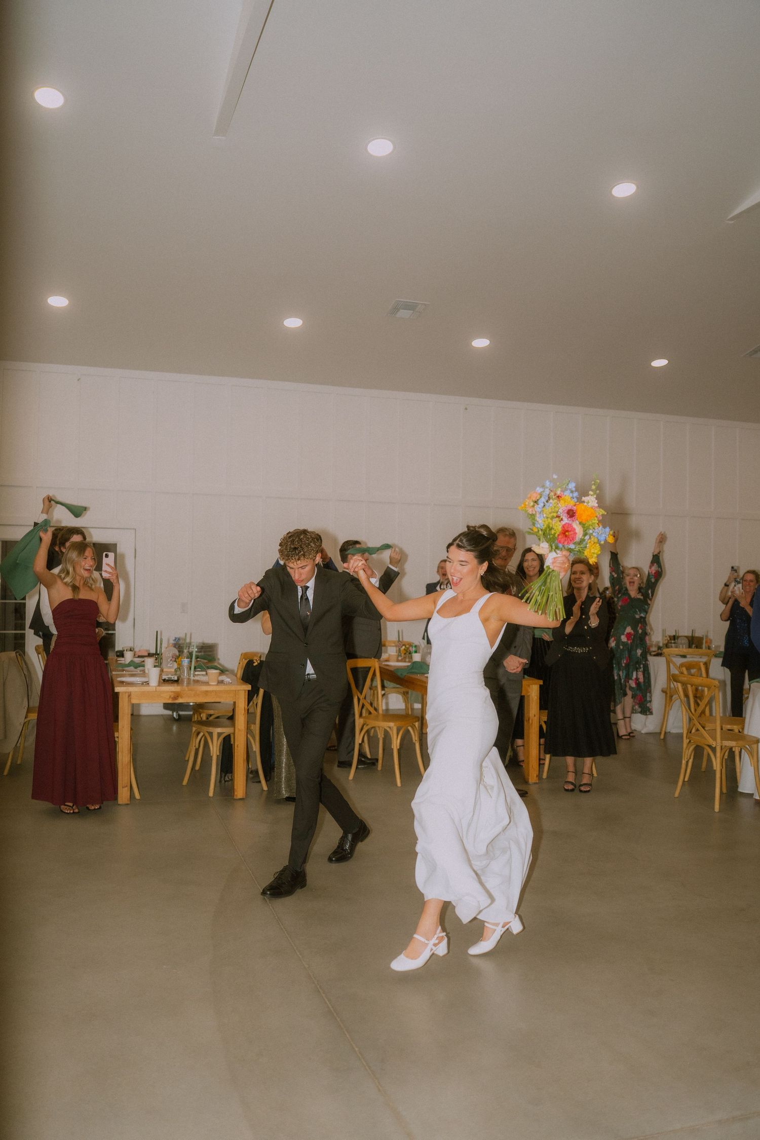 Wedding reception dancing in a bright indoor venue with wooden chairs.