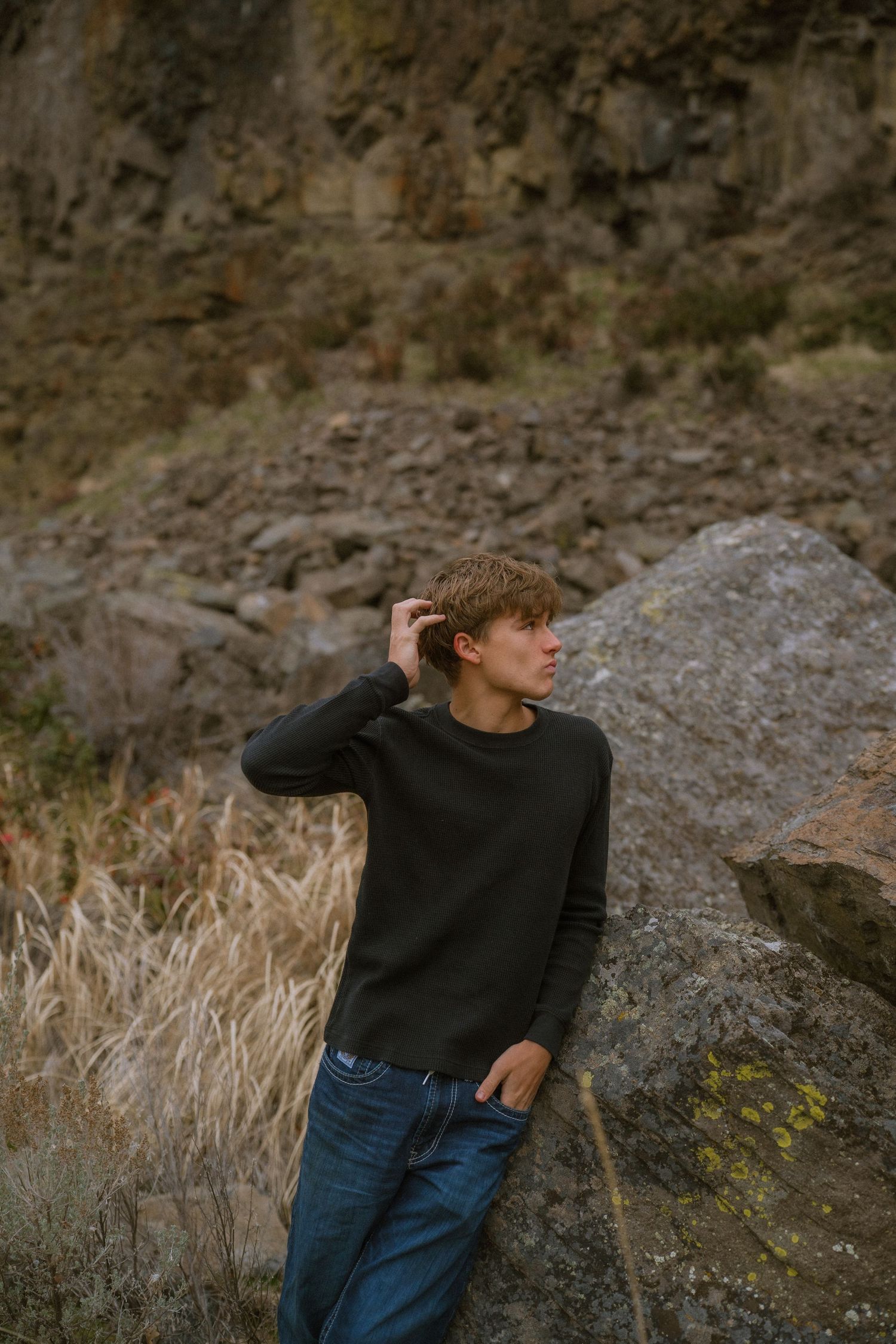 Person in black sweater and jeans poses against rocky cliff backdrop with dried grass and natural scenery.