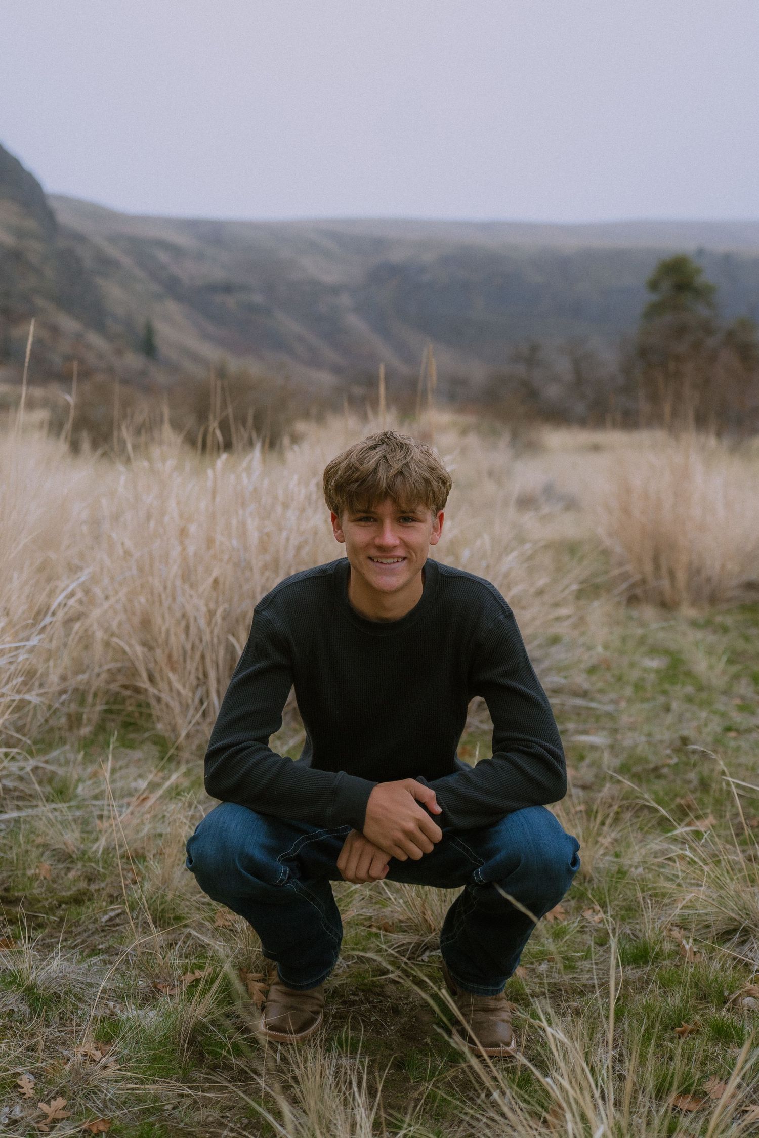 A person wearing dark clothing sits in tall grass on a hillside with mountains visible in the background.