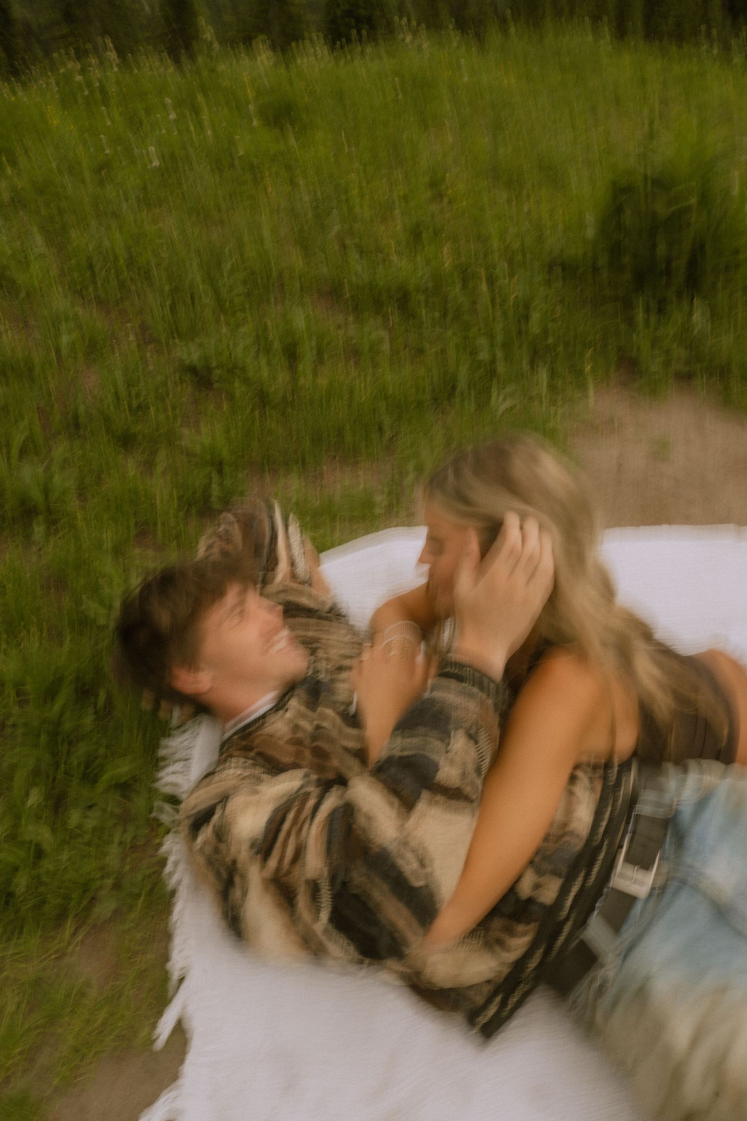 Two people share an intimate moment lying in tall grass with mountains in the background.