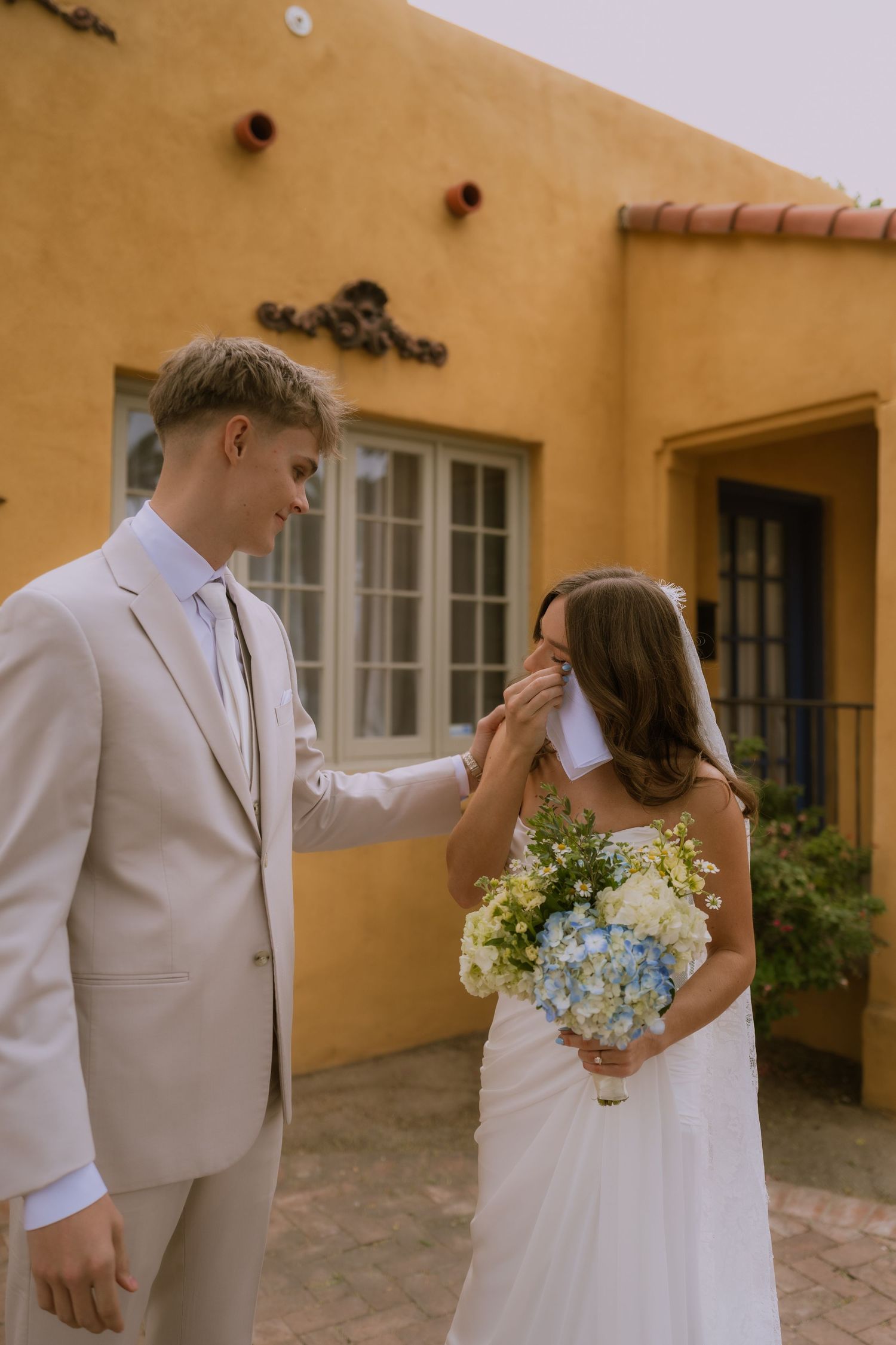 A wedding couple shares an emotional moment in front of a Spanish-style stucco building with a beautiful bouquet of flowers.