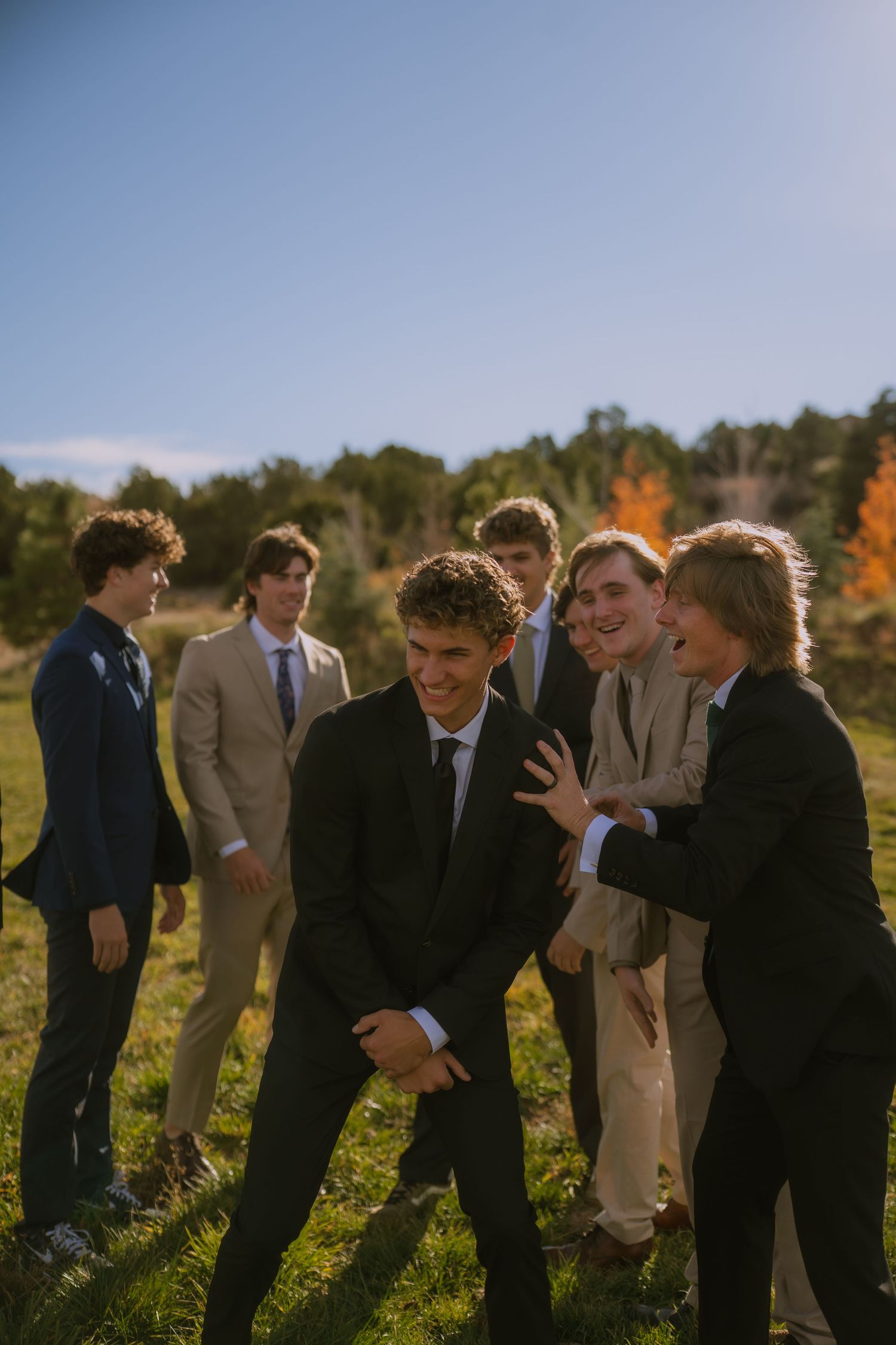 Groomsmen in suits laughing and joking together in a sunny outdoor field during a wedding photoshoot.