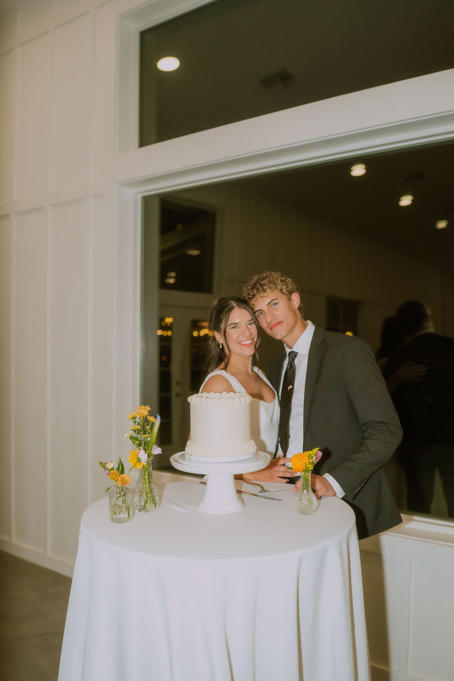 Wedding couple standing at white cake table with sunflowers in vases.