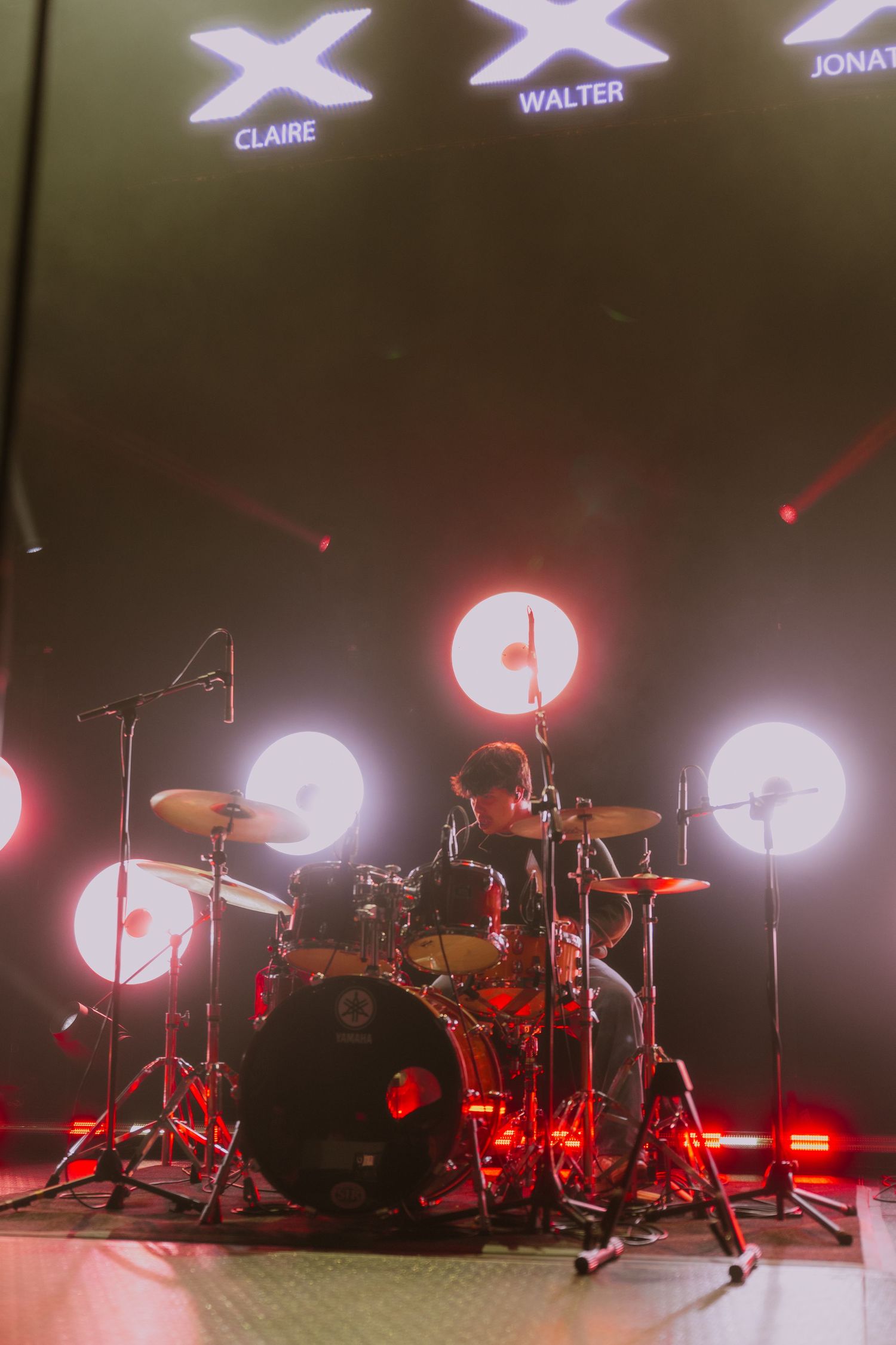 Drum kit setup on stage with red lighting and illuminated name displays visible in background.