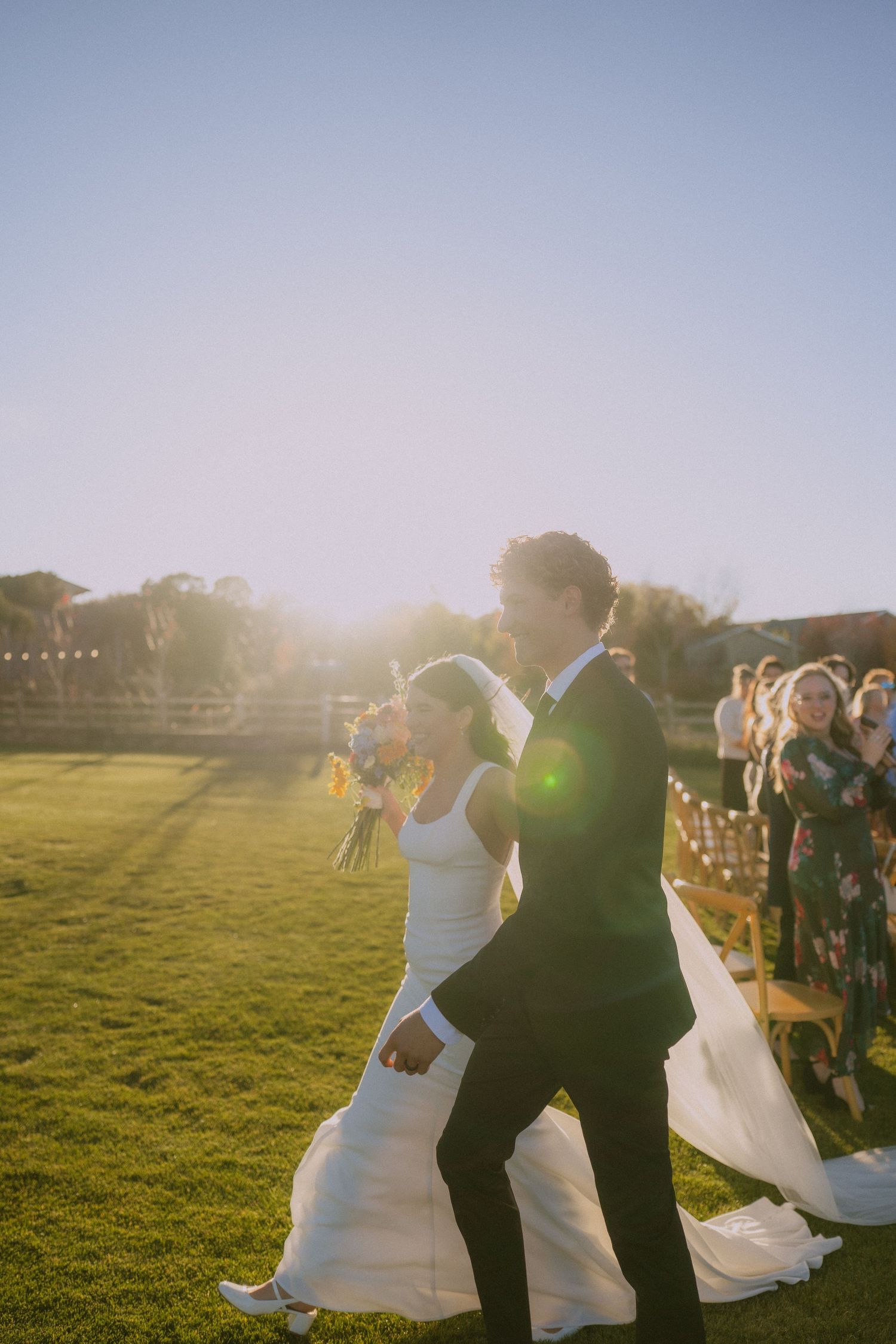 Wedding guests gather on a sunlit lawn during an outdoor celebration.