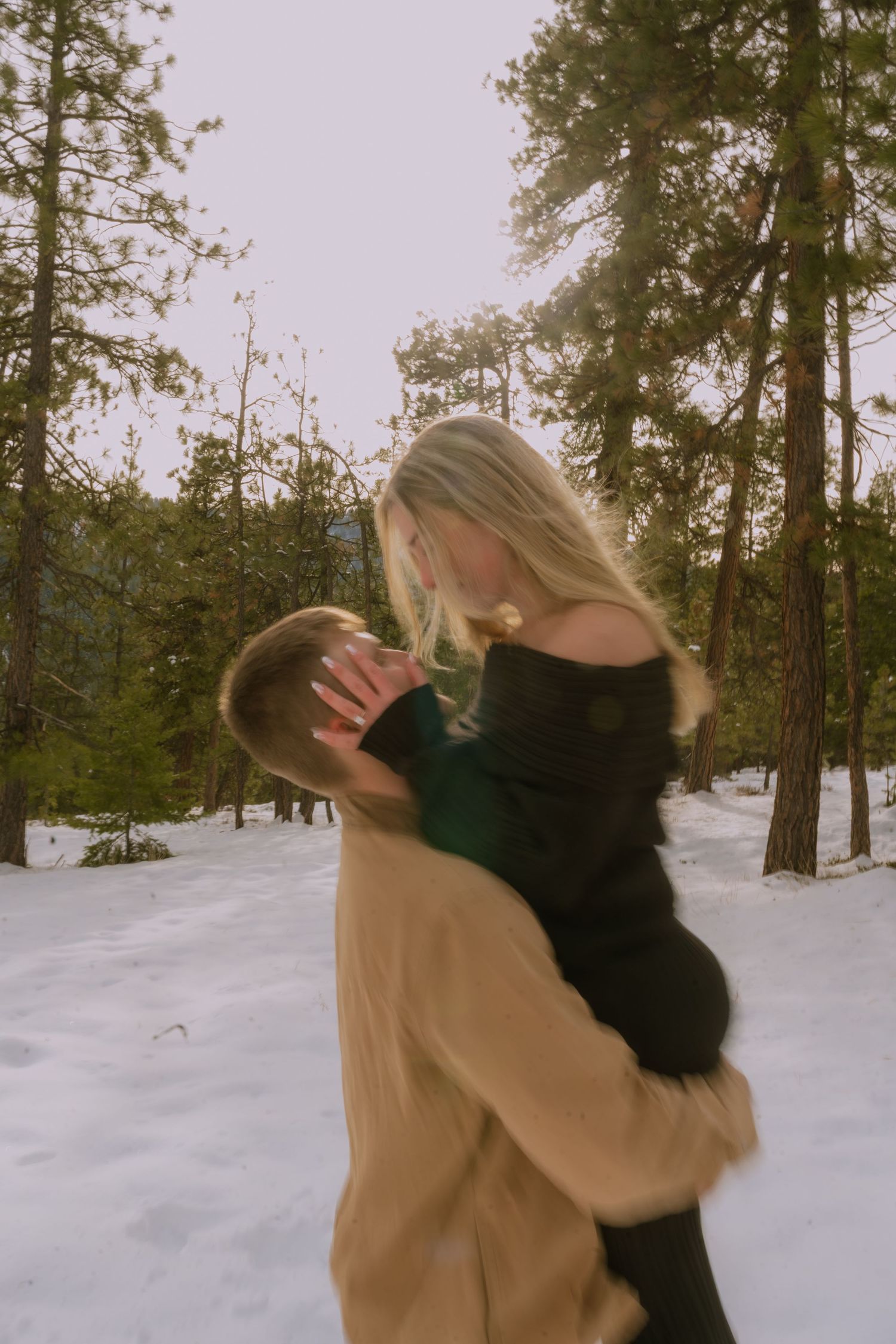 Two people in winter coats embrace in a snowy forest landscape with tall pine trees in the background.