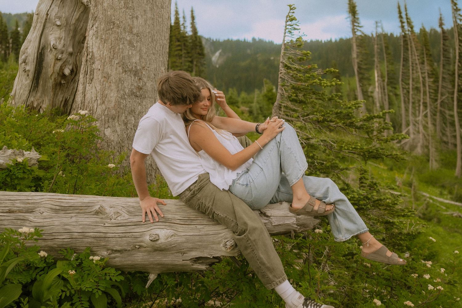 A couple relaxes on a fallen log surrounded by evergreen trees in a mountain landscape.