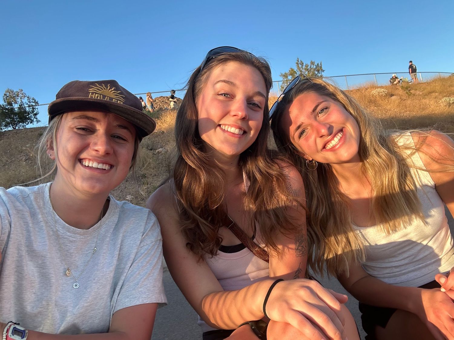 A group of friends taking selfies together outdoors during golden hour with scenic landscapes in the background.