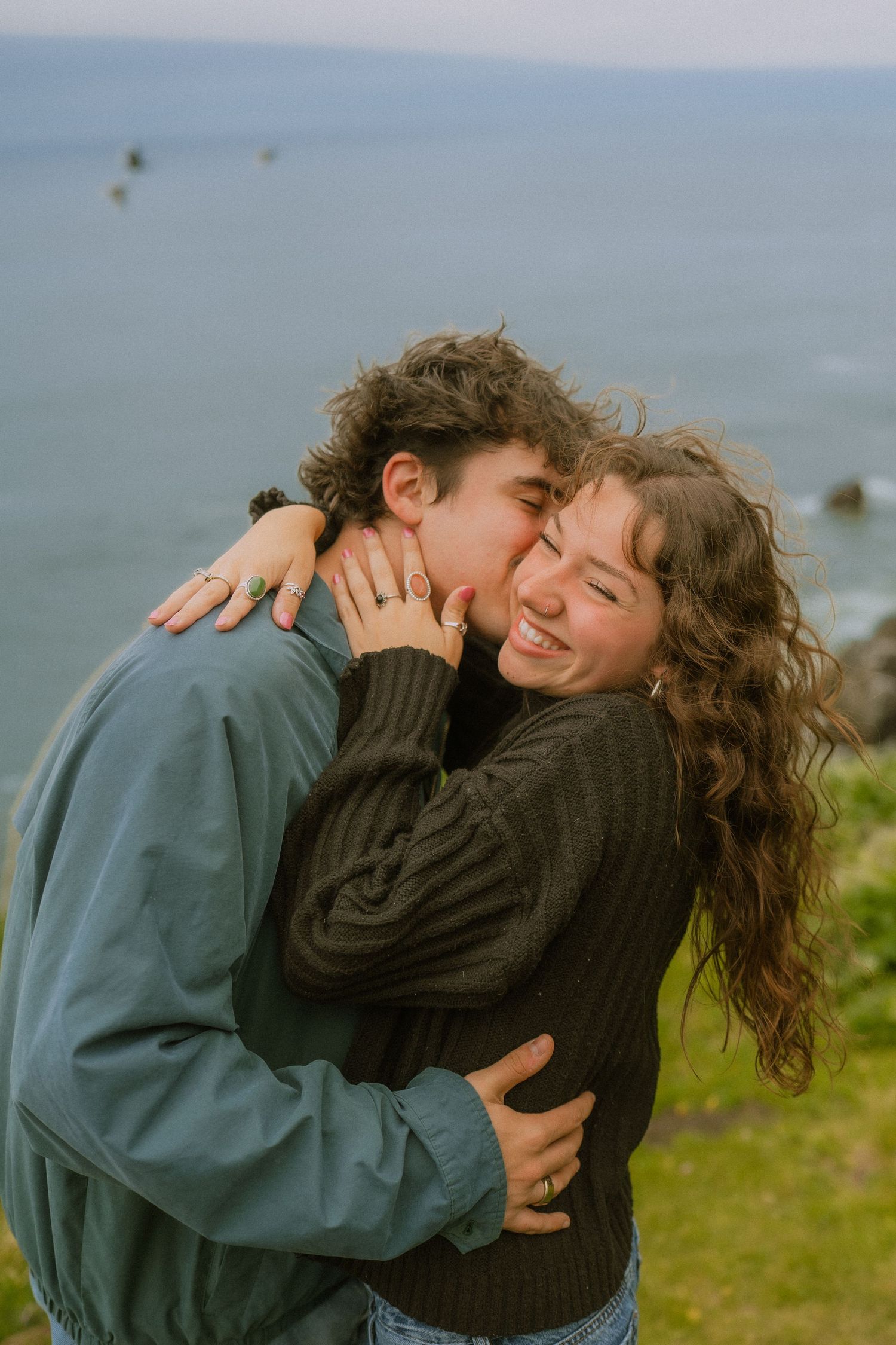 A couple shares a tender embrace while laughing together on a coastal cliff overlooking the ocean on an overcast day.