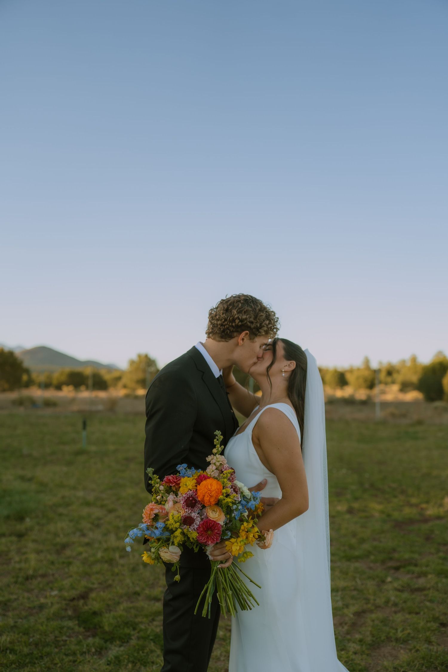 A couple shares a romantic kiss in a desert field at sunset while holding a vibrant wildflower bouquet.