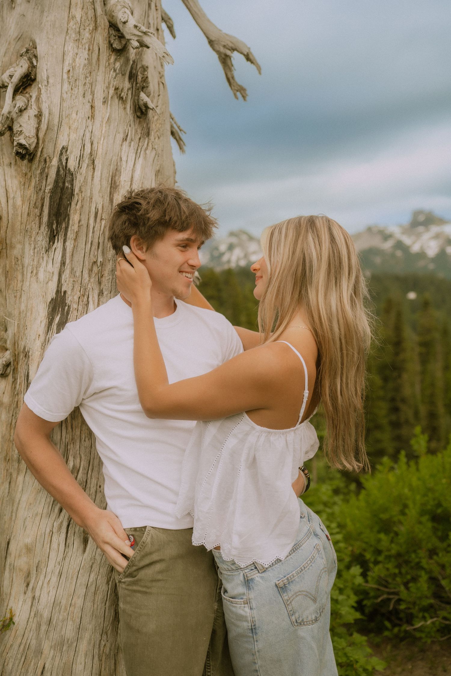 A couple in casual white shirts and denim embraces playfully against a rustic wooden backdrop in nature.