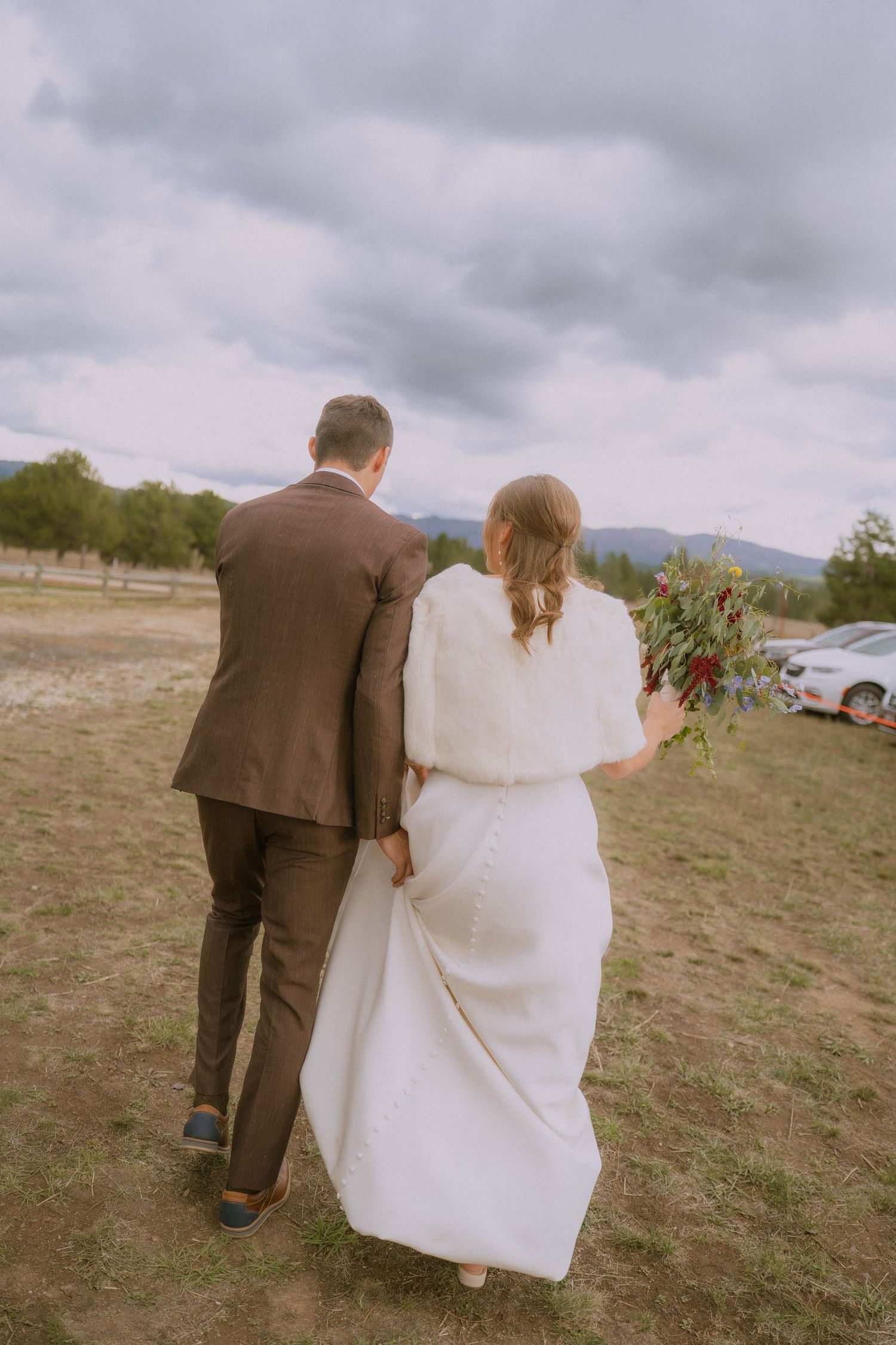 A couple in wedding attire walking together on a grassy field under cloudy skies, with mountains in the distance.