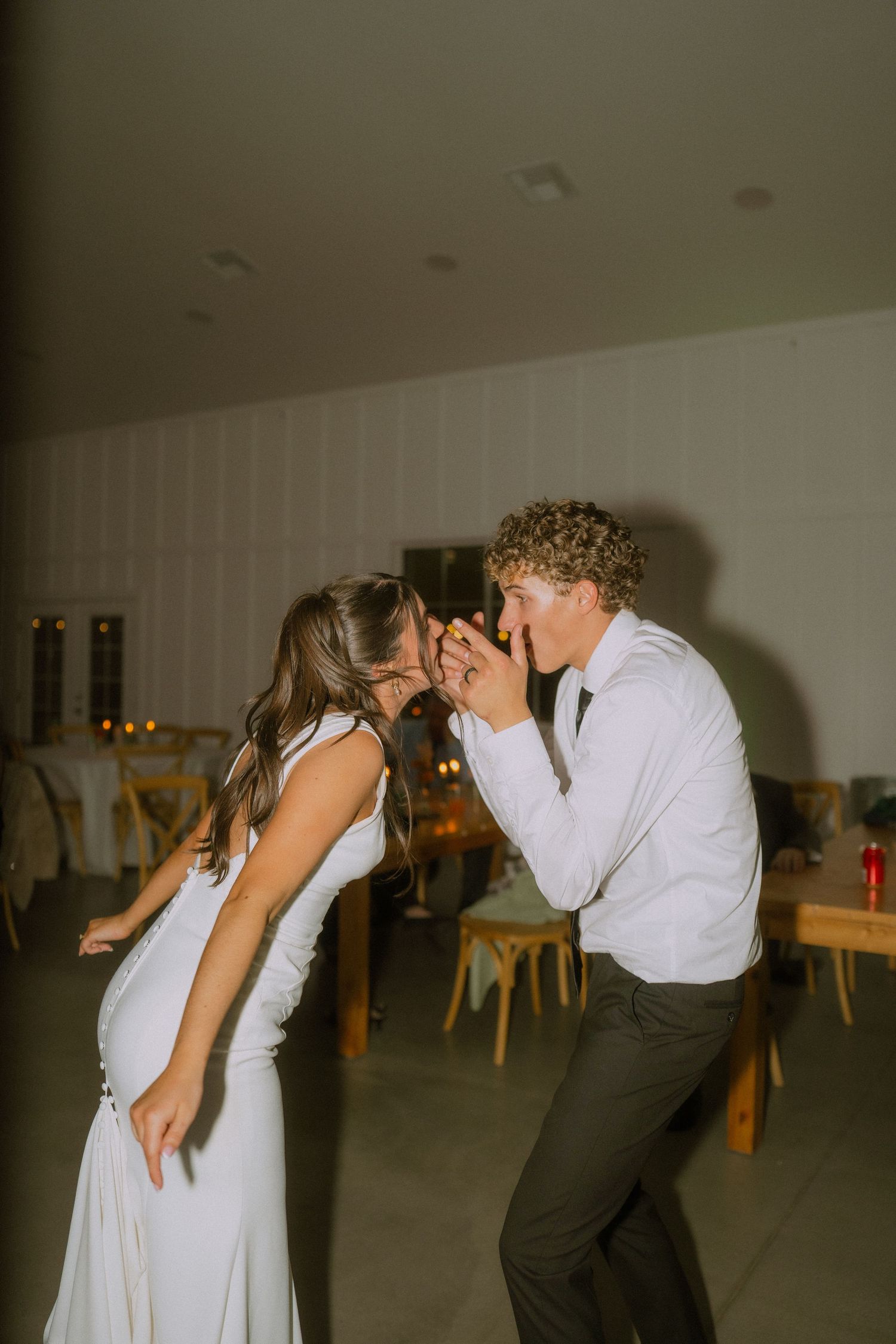 Couples dance and celebrate in a rustic indoor reception venue with wooden tables and dim lighting.