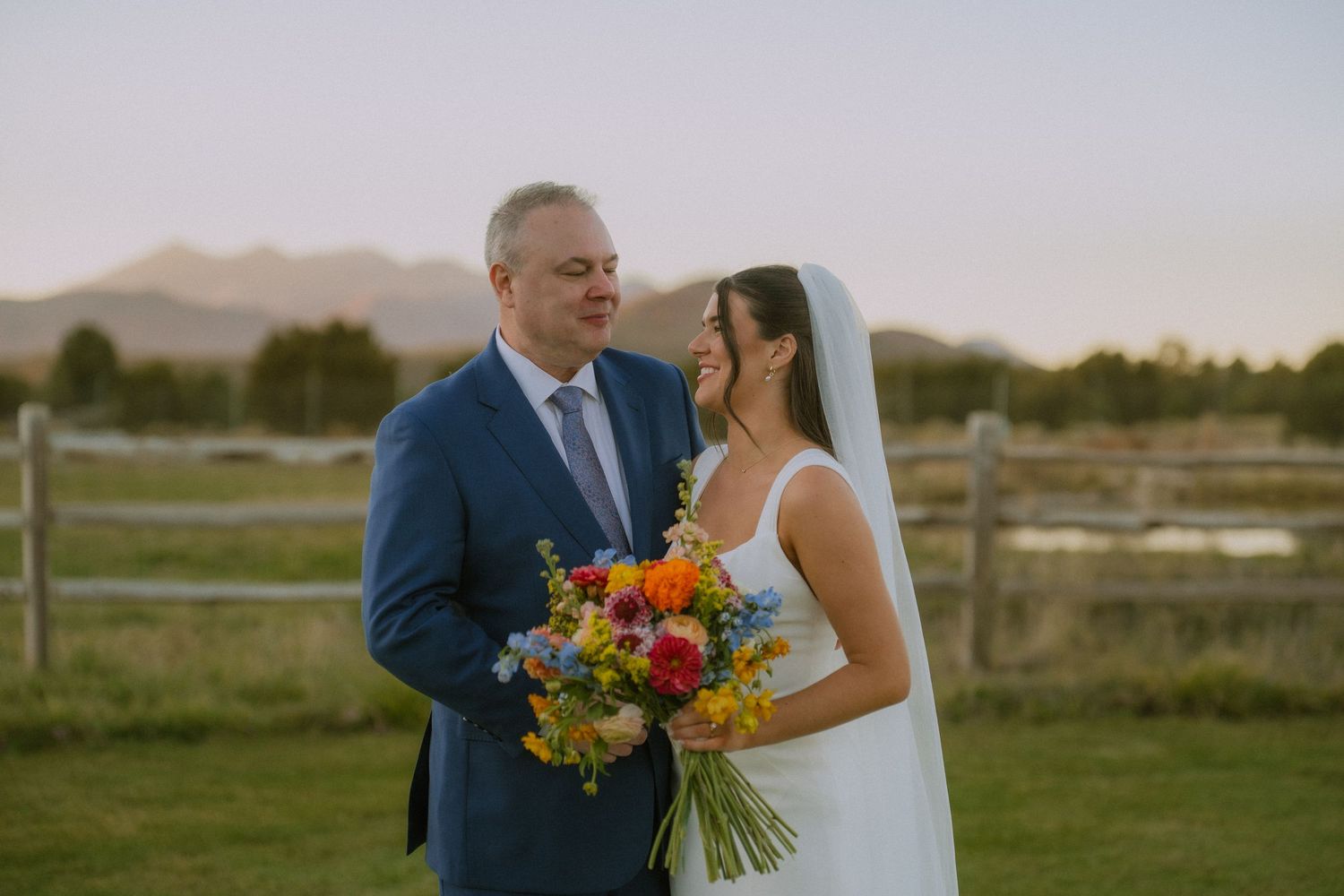Bride in white dress holding colorful bouquet poses outdoors with rustic fence background.
