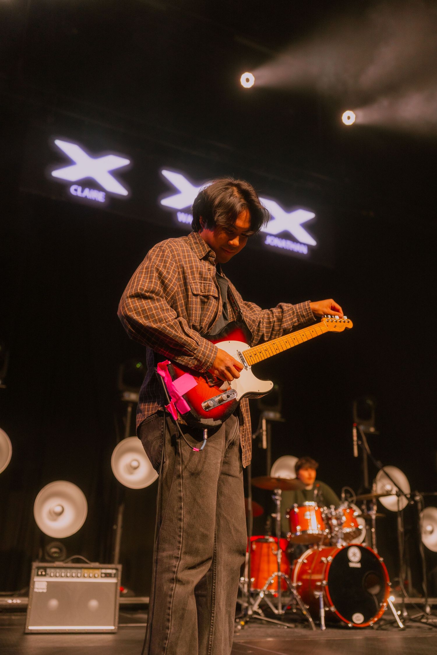 Guitarist performs on stage with red guitar in front of drum kit during live concert with dramatic lighting.