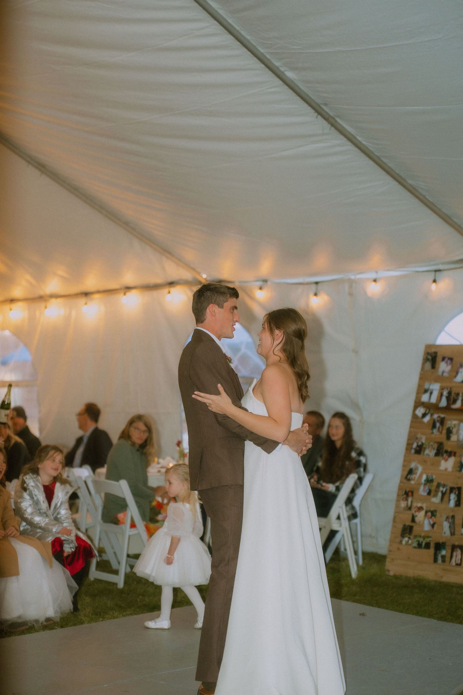 A couple shares their first dance under a white tent decorated with string lights at their wedding reception.