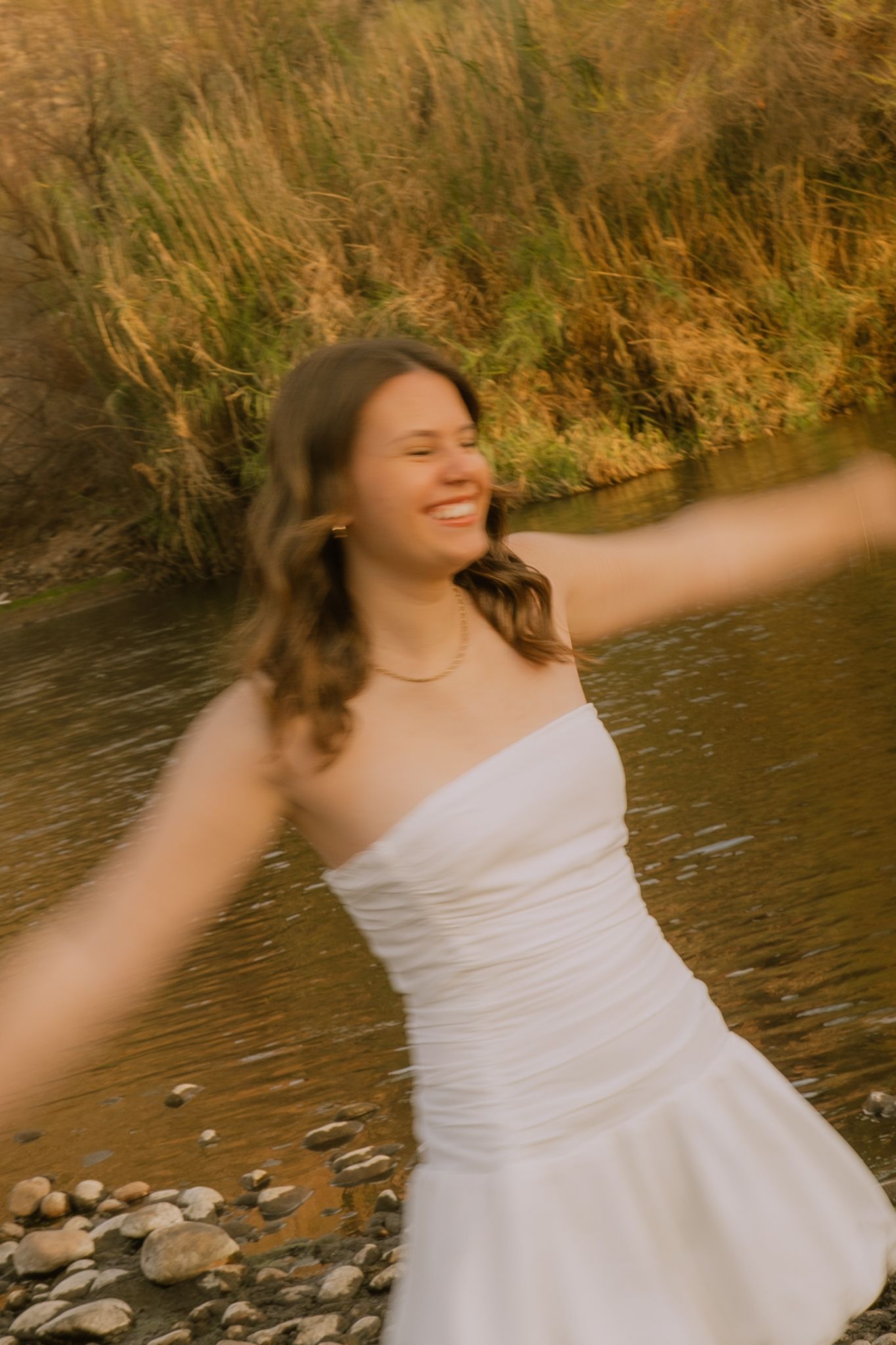 A person in a white dress spins joyfully by a river with golden grass in the background during sunset.