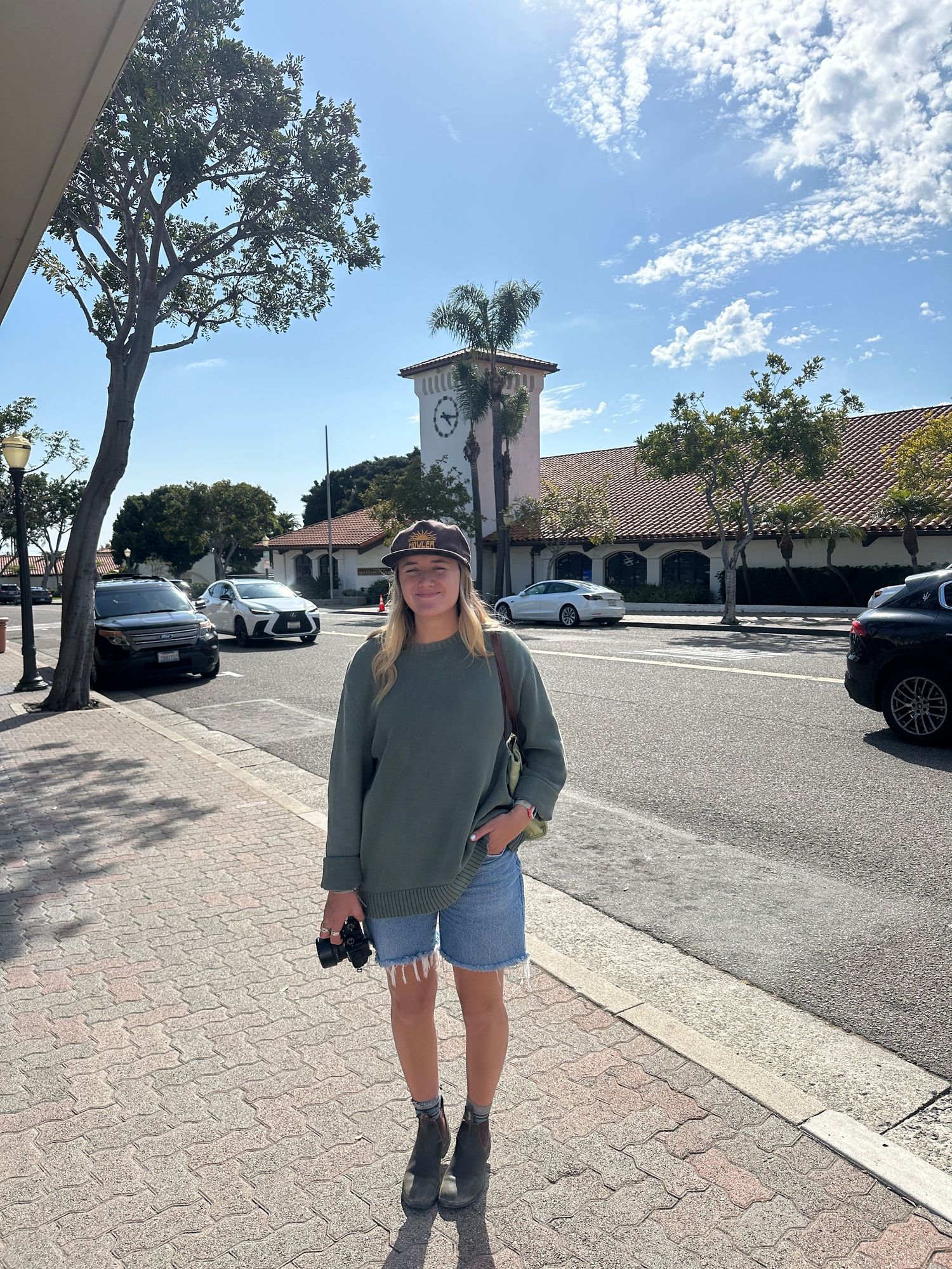 A person in casual attire walking along a sunny brick-paved sidewalk near Spanish-style buildings and parked cars.