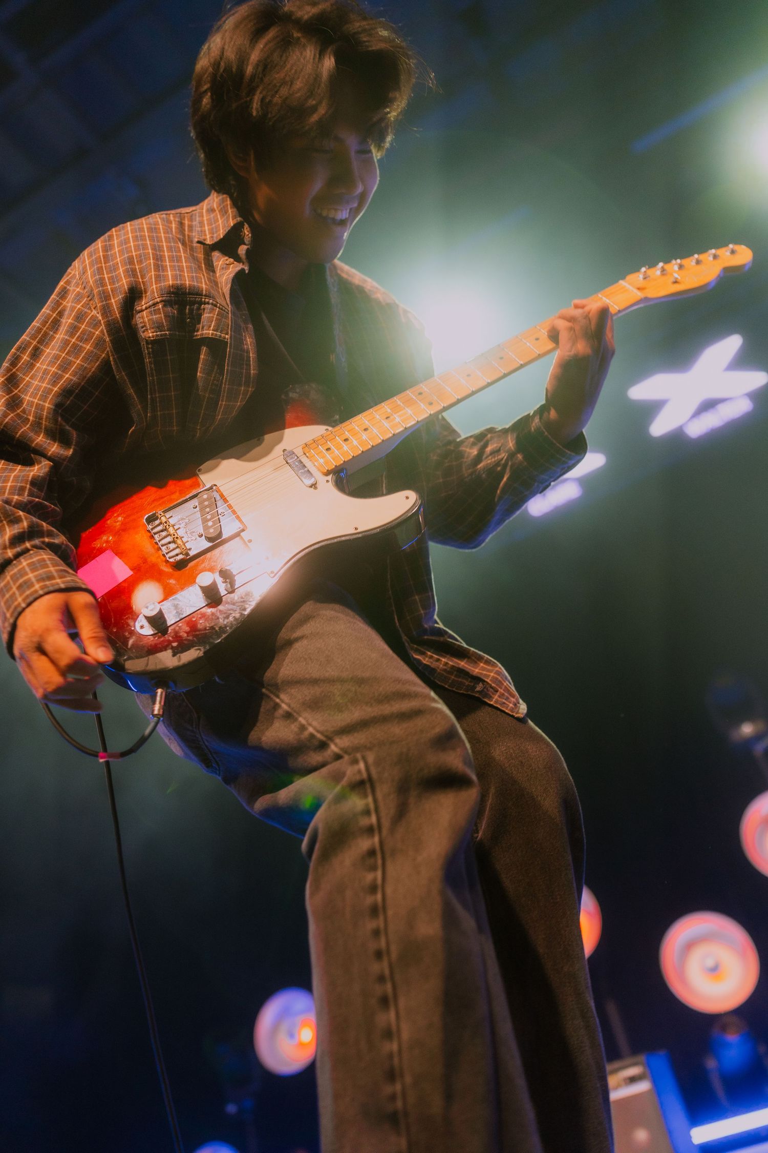 A guitarist performs on stage with an orange electric guitar against a dark background with blue stage lighting.