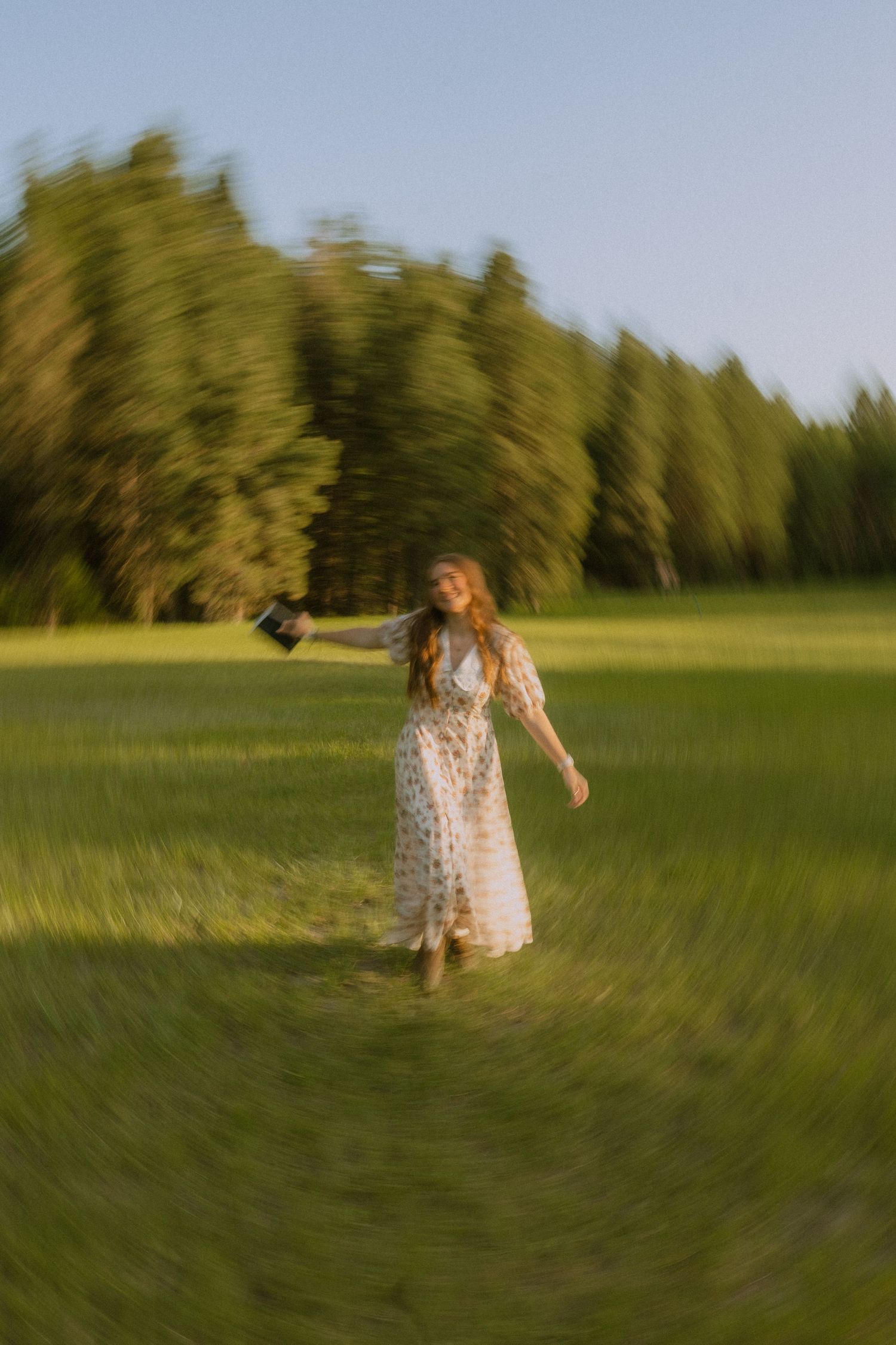 Motion-blurred figure in a white floral dress spins through a sunlit green field at golden hour.