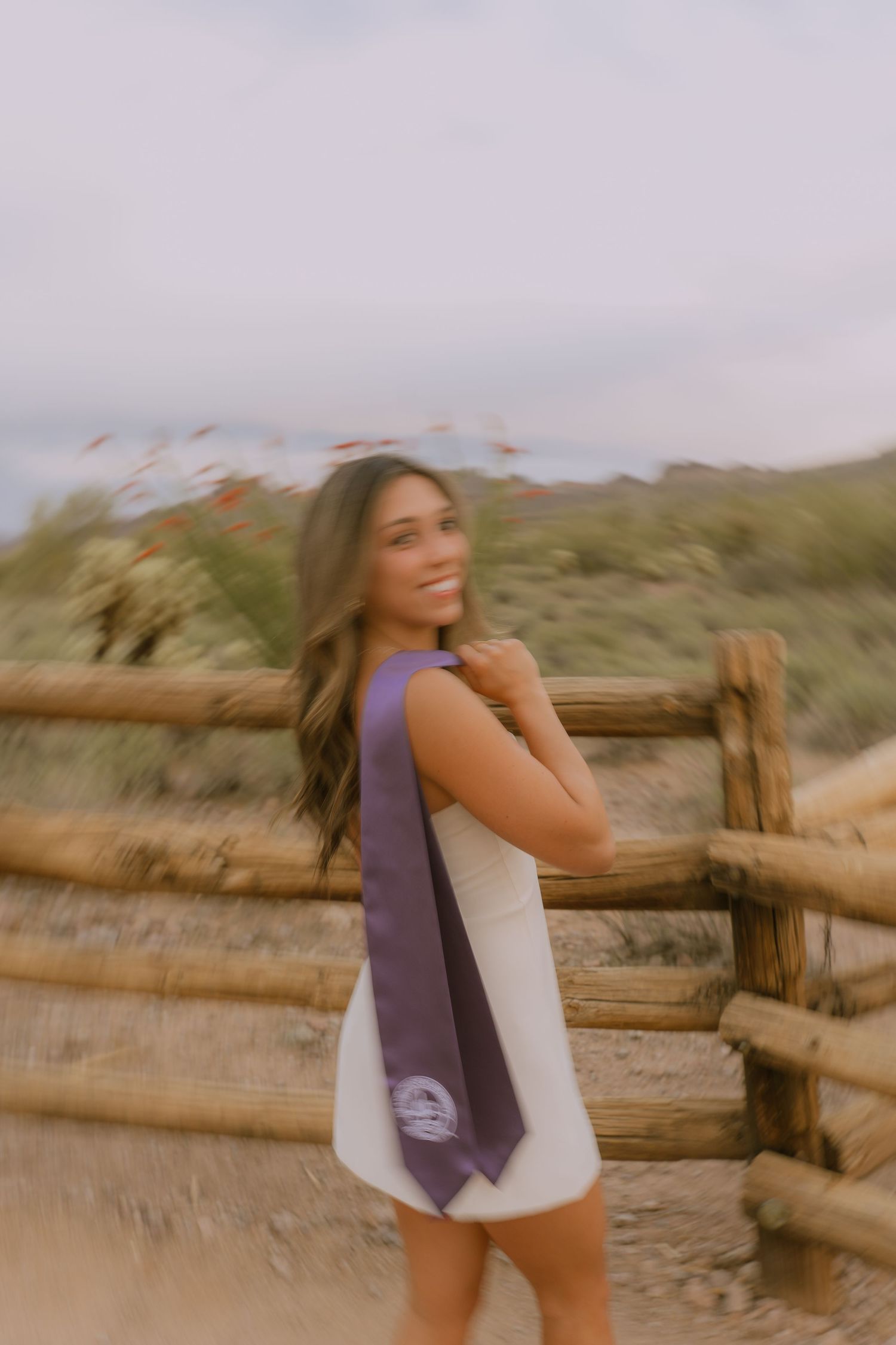 A person in a white top and purple vest poses against a rustic wooden fence in a desert landscape at sunset.