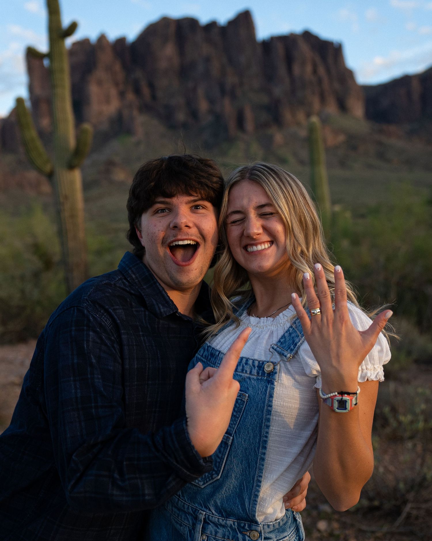 A couple shows off their engagement rings while posing in front of desert mountains and saguaro cacti at sunset.