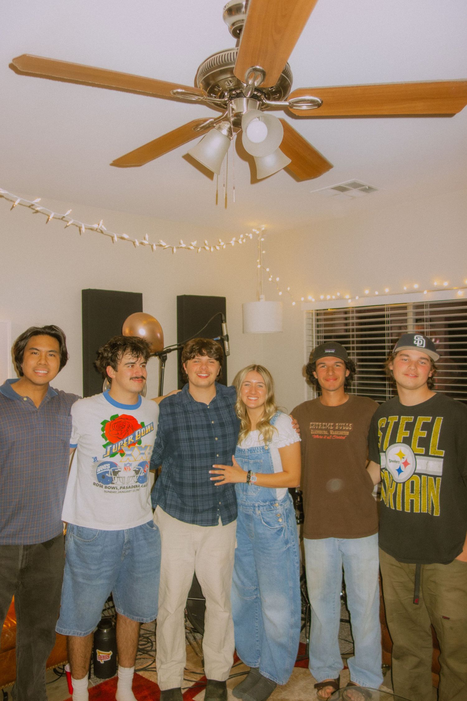 A group of people posing together in casual clothing under string lights and ceiling fans at an indoor gathering.