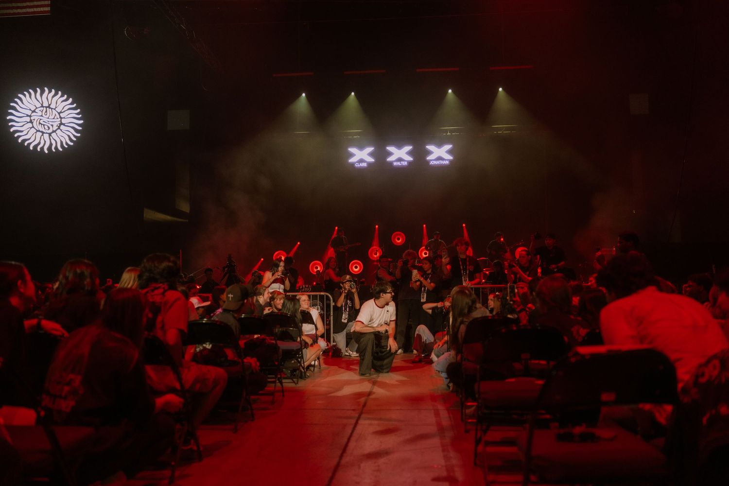 Concert stage lit with dramatic red lighting shows silhouetted performers and audience members in a dark venue.