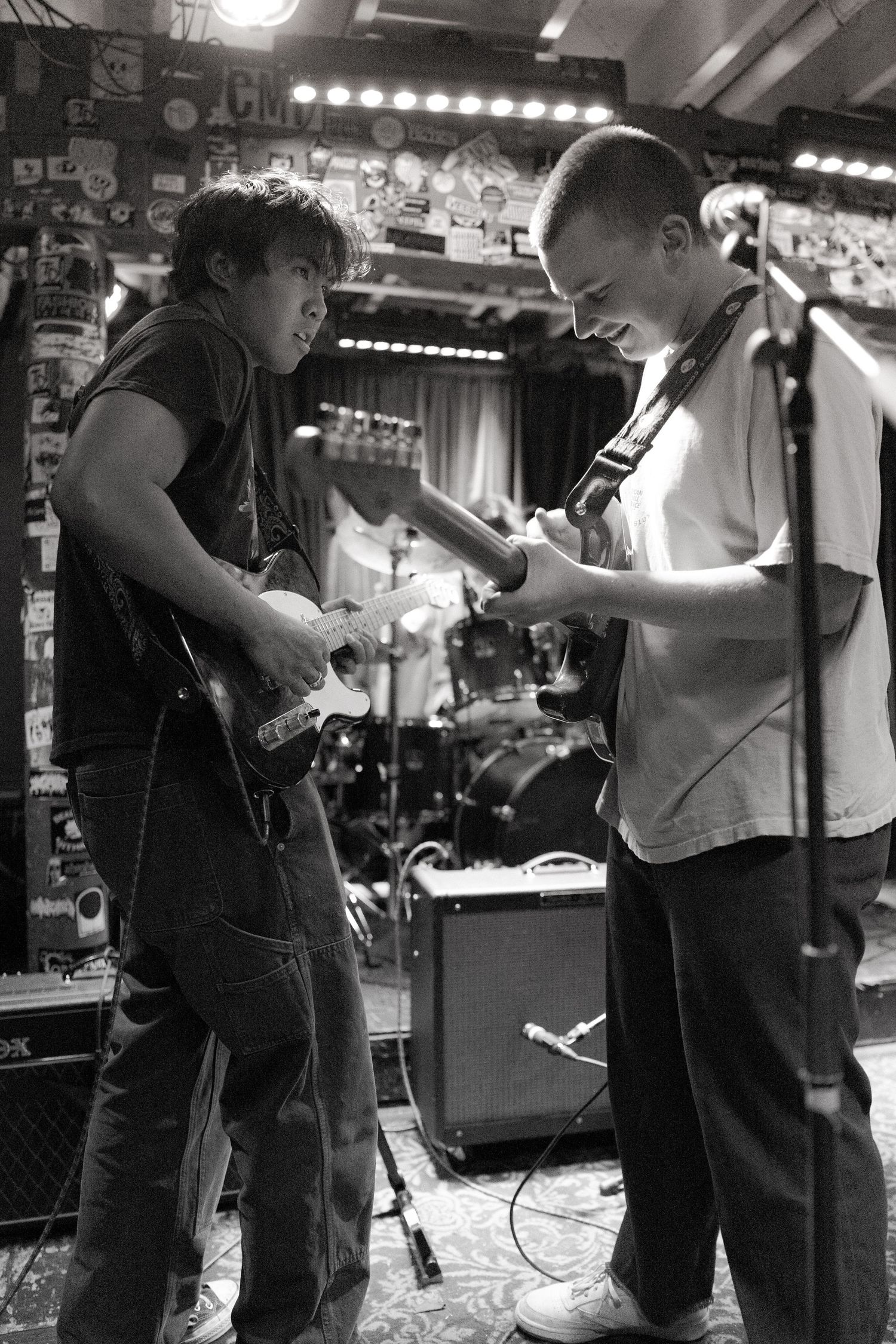 Musicians perform on stage in a dimly lit venue with equipment and lighting visible in the background.