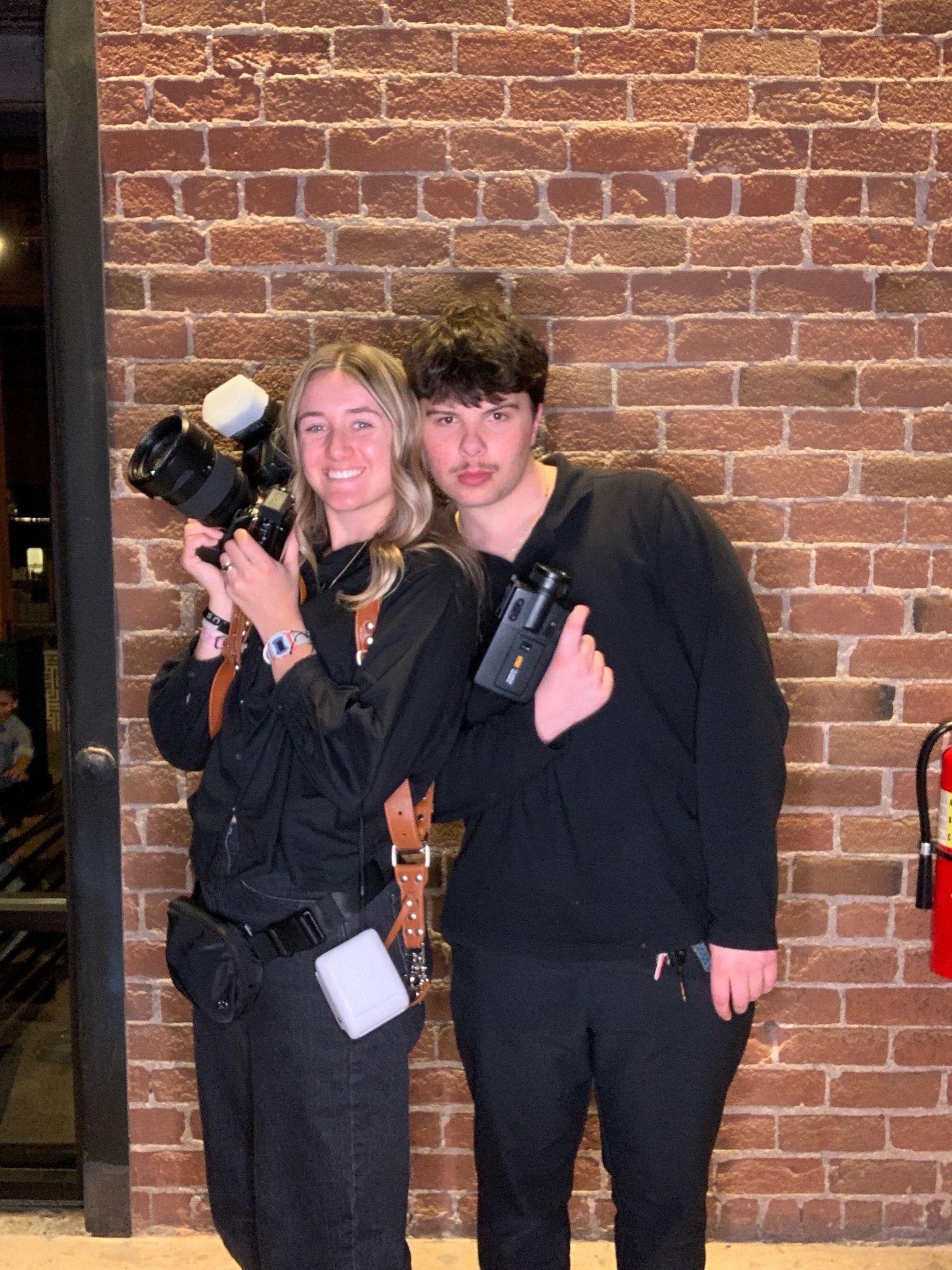 Two photographers with professional cameras pose together against a brick wall in matching black outfits.