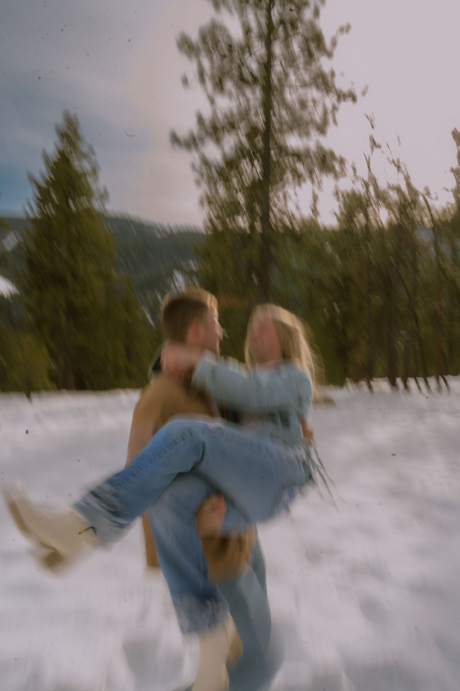 A couple plays in the snow together on a winter day with evergreen trees in the background.