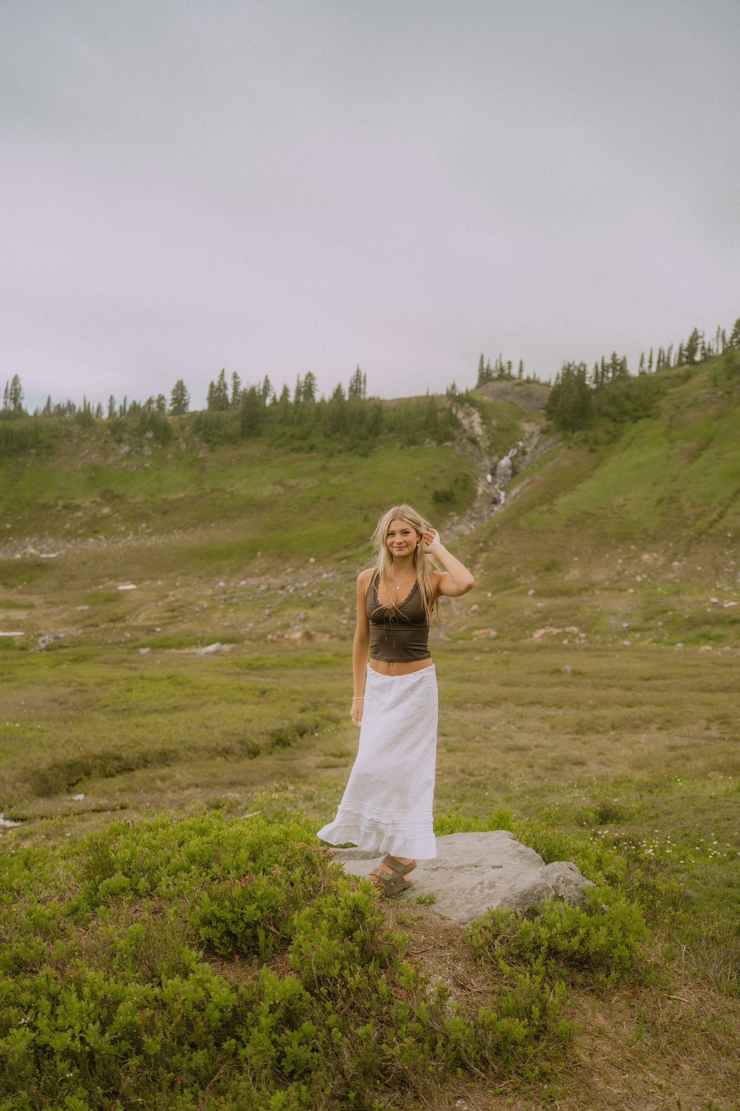 A figure in a flowing white skirt stands on a rocky outcrop surrounded by alpine meadow and evergreen trees.