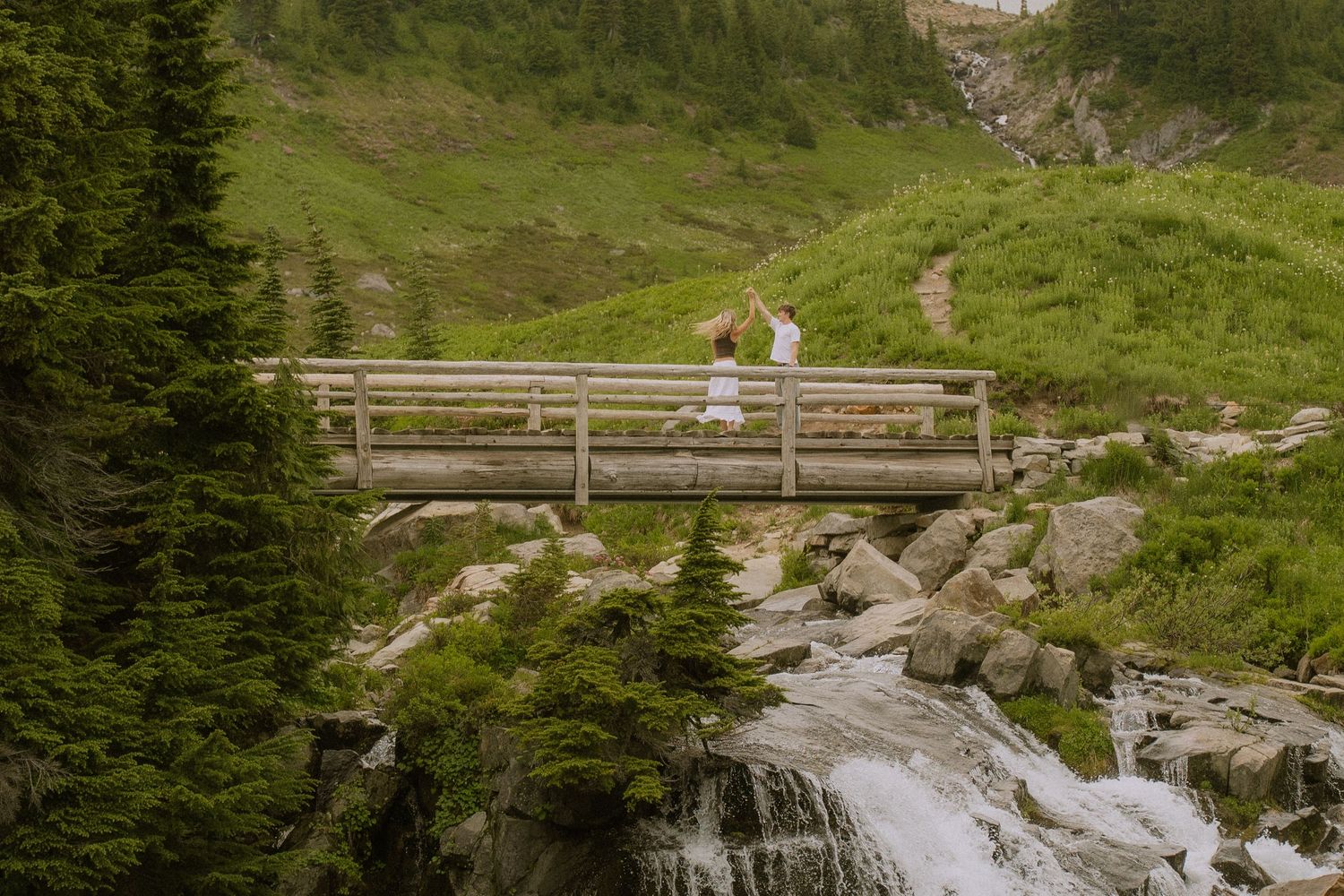 A scenic mountain waterfall cascades down rocky terrain surrounded by lush green forest and hiking trails.