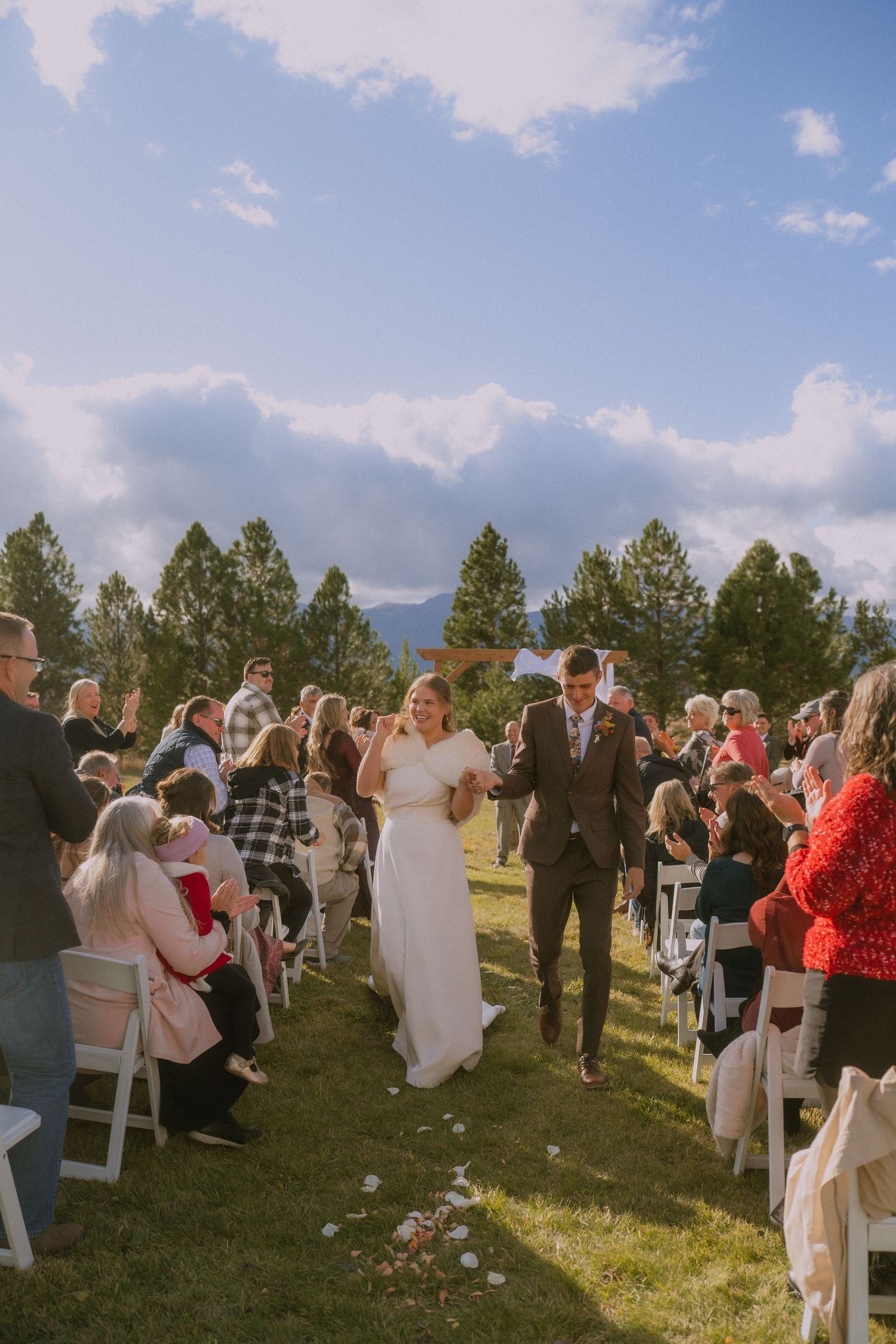 Newlyweds walk down the aisle at an outdoor wedding ceremony surrounded by guests and pine trees on a sunny day.