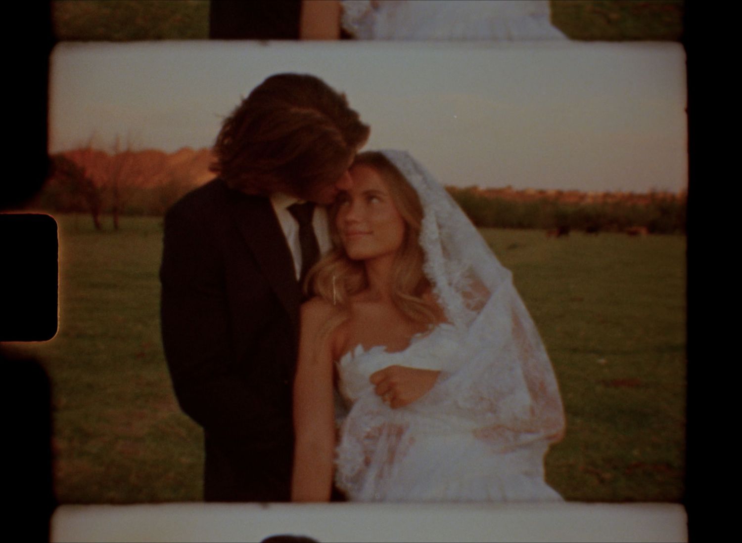A bride in a white lace gown and veil stands with a groom in a black suit outdoors at a wedding.